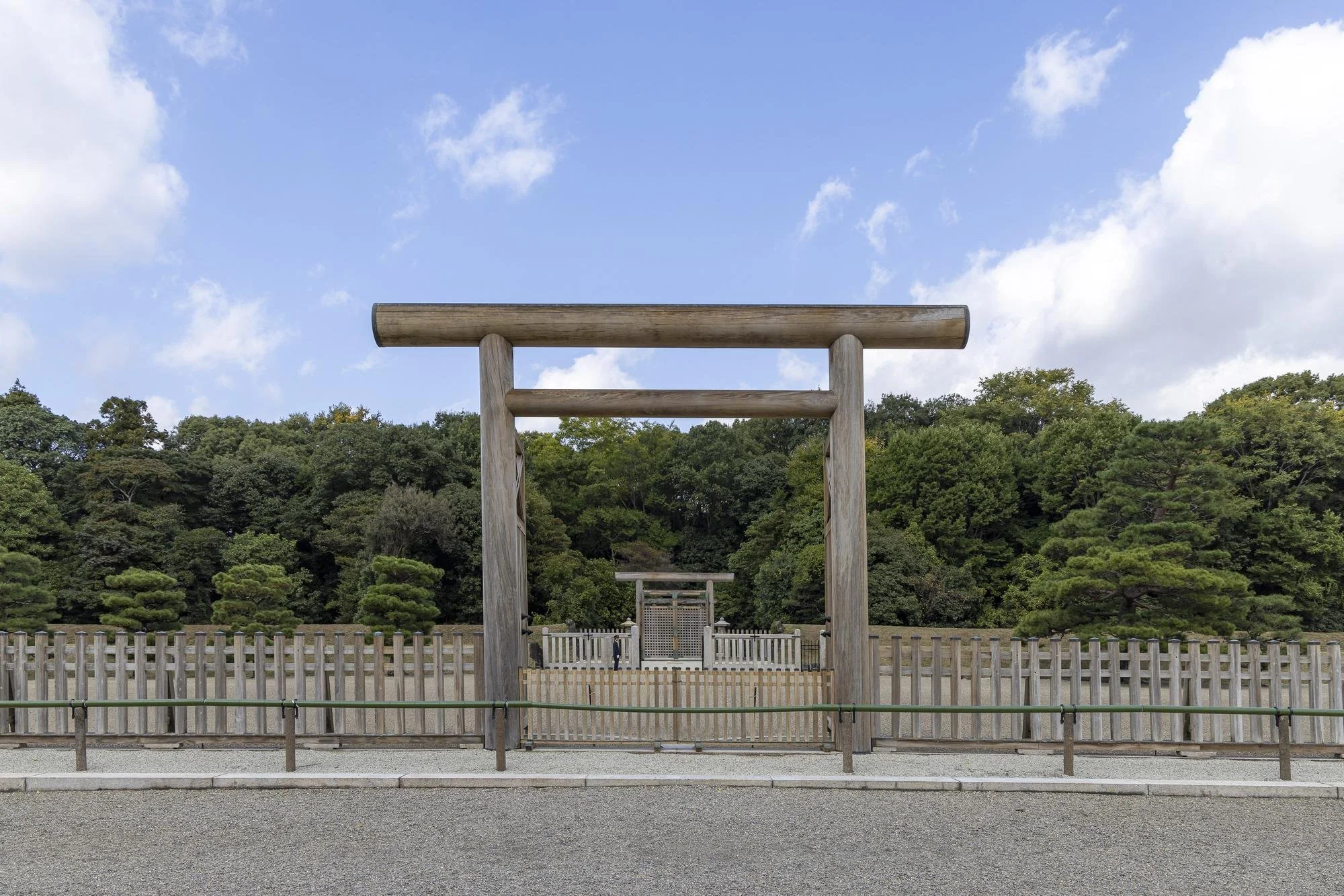 A traditional Japanese torii gate made of wood in front of a pathway leading into a shrine, with a forested area and a partly cloudy sky in the background.