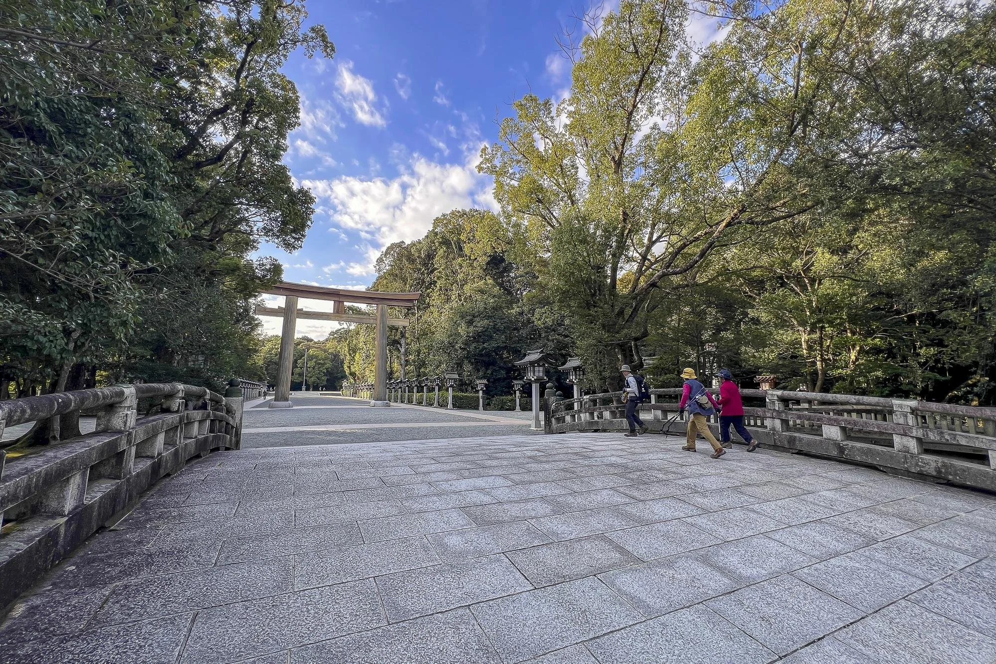 Four people walking on a stone pathway near a traditional Japanese torii gate, surrounded by lush green trees under a blue sky with scattered clouds.