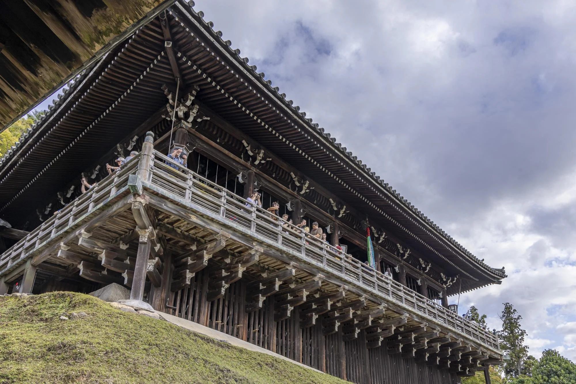 Traditional Japanese wooden temple with people on the balcony, against cloudy sky and trees.
