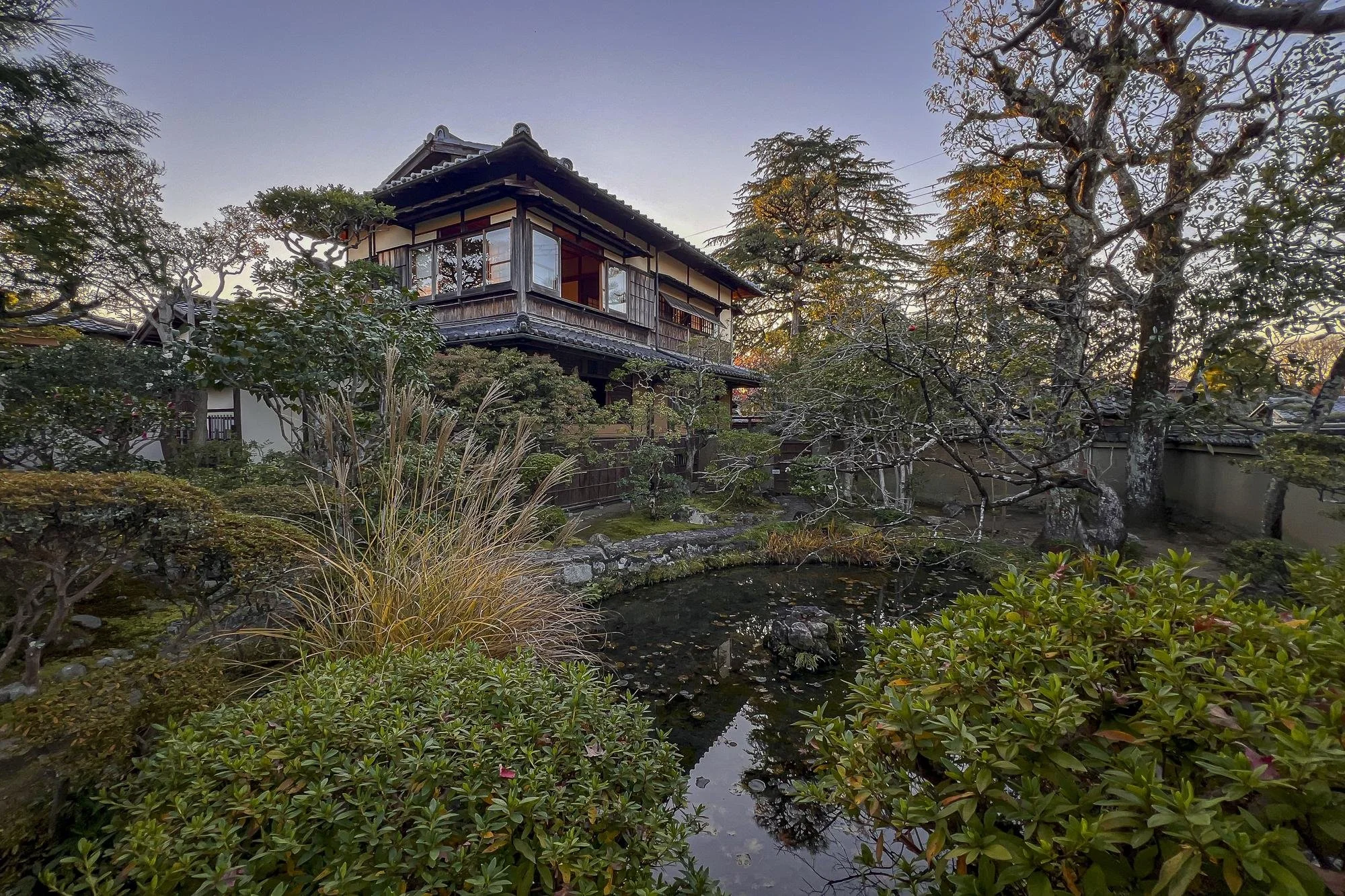 Traditional Japanese garden with a pond, lush greenery, trees, and a wooden house with sliding doors and balcony in the background.