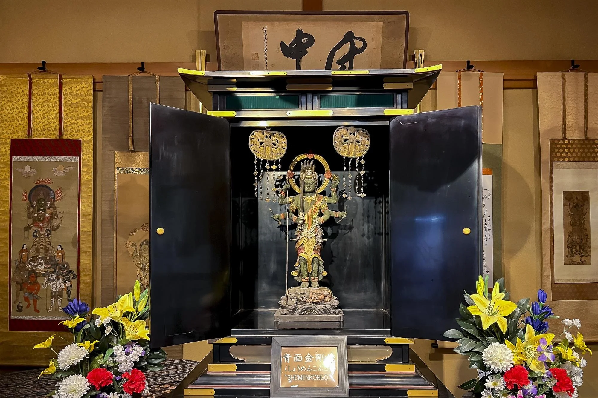 A religious altar with a statue of a multi-armed deity inside a black wooden cabinet. The altar is decorated with colorful flowers at the base and has a sign with Japanese writing and the title 'Shomenkongo' below the statue.