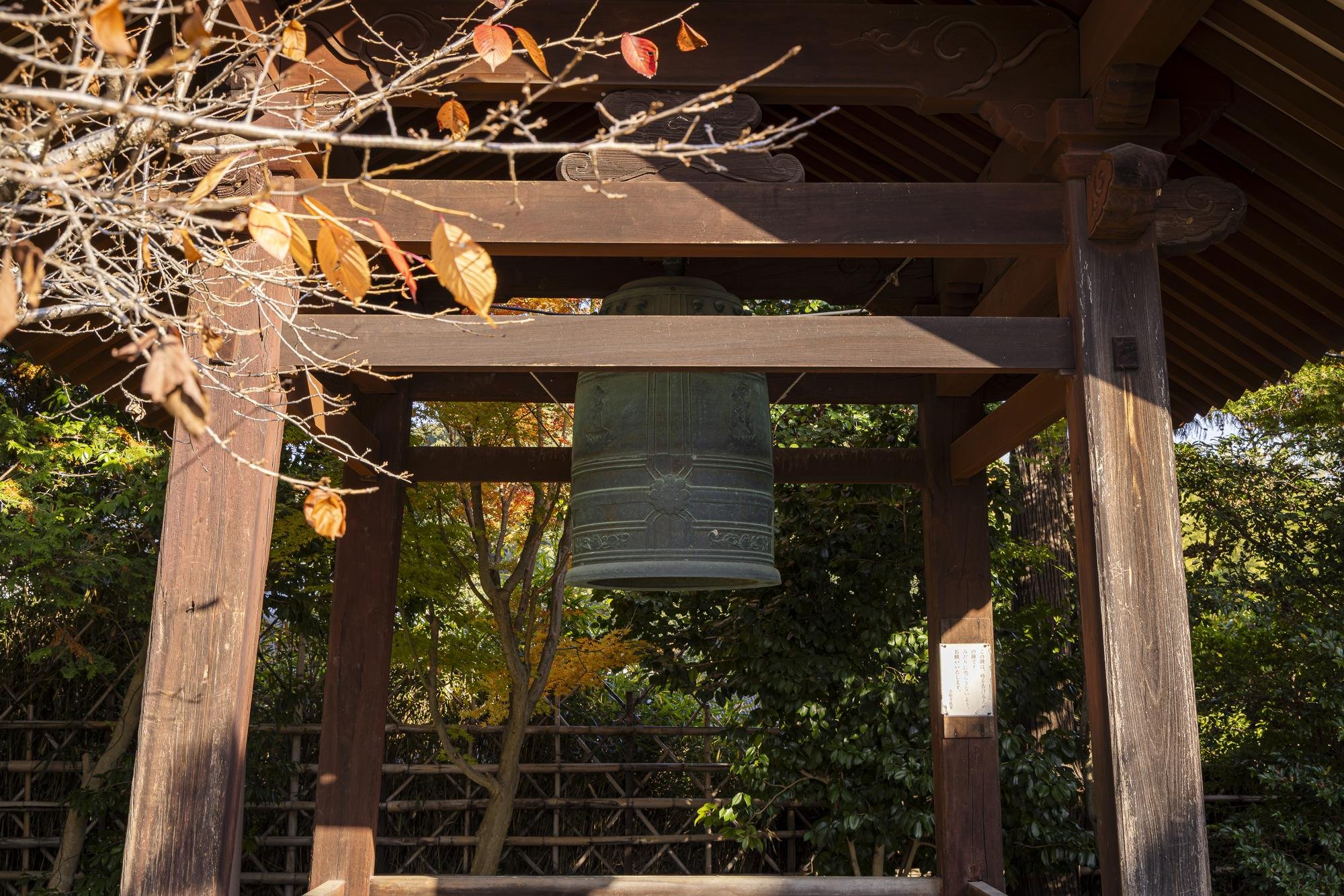 A traditional Japanese temple bell hanging under a wooden structure with autumn leaves in the foreground.