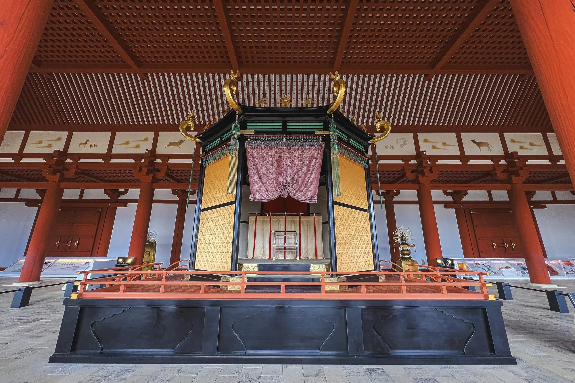 Interior of a traditional Japanese shrine with a central elevated structure, decorated with gold accents, pink curtains, and surrounded by red wooden pillars and railings.
