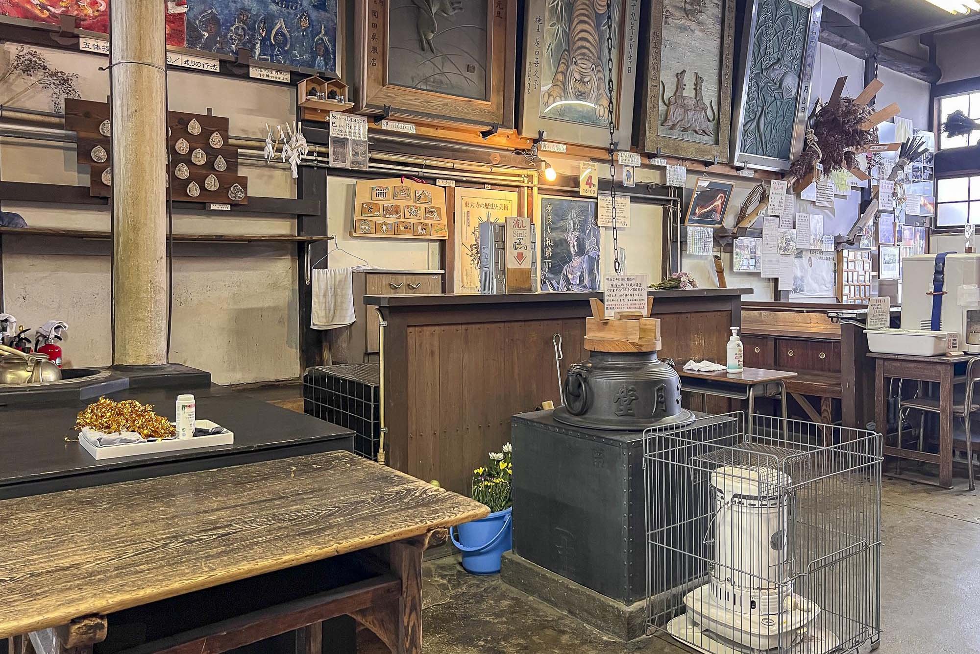 Interior of a Japanese restaurant or shop with various traditional artwork and decorations on the walls, a wooden table in the foreground, and a counter area with a sign, sanitizer bottle, and utensils. There is steam and a covered kettle or stove in