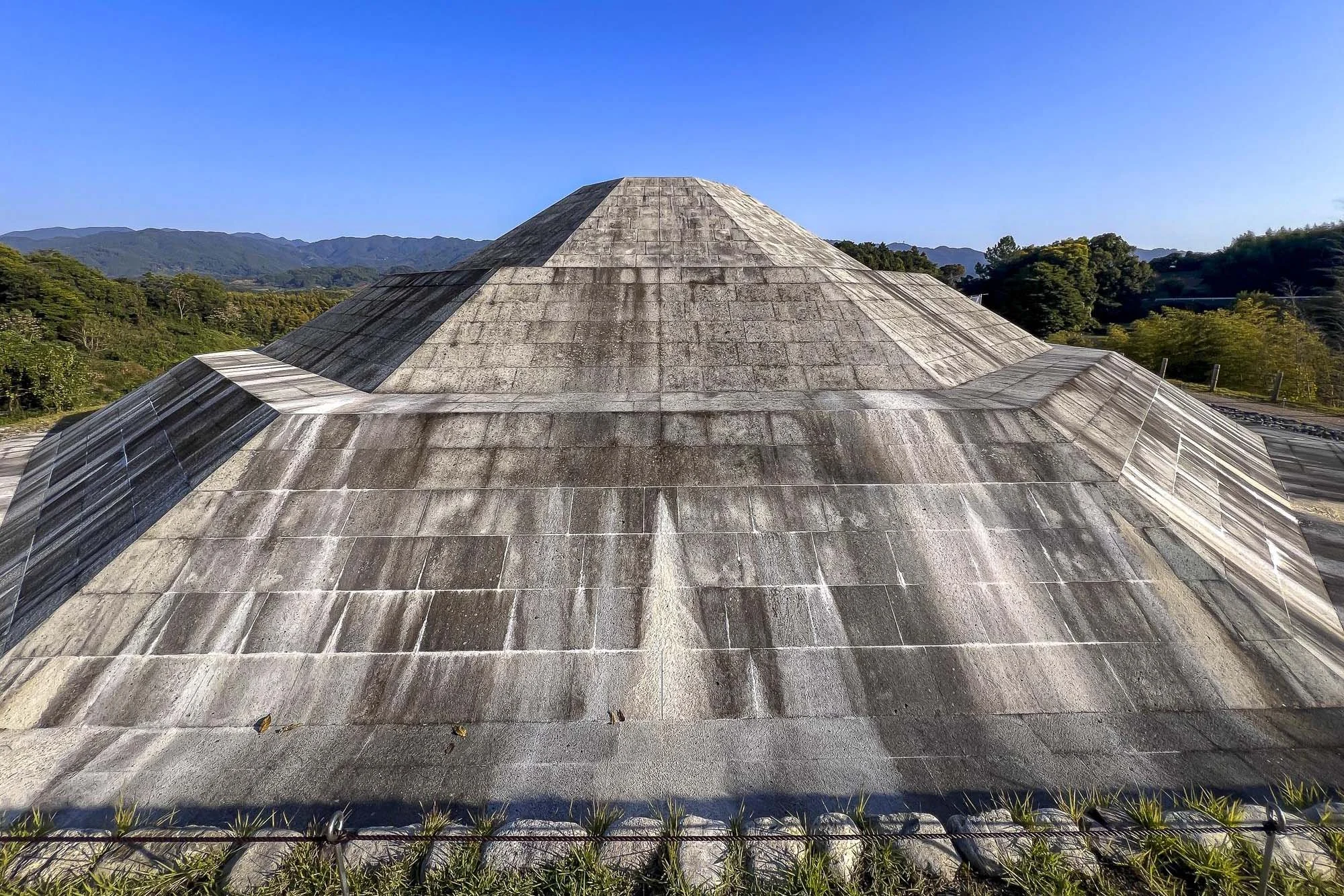 Large concrete domed structure with a stepped design, surrounded by a natural landscape of trees and mountains in the background under a clear blue sky.