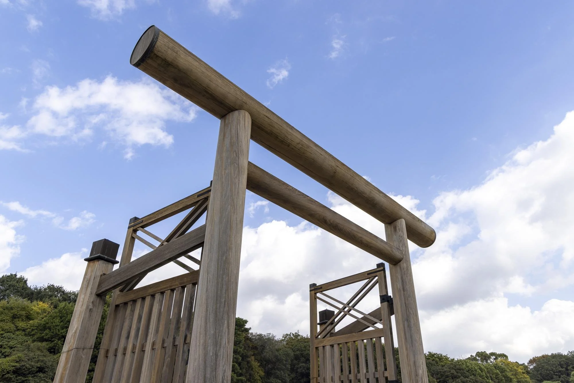 A wooden frame structure resembling a traditional Japanese torii gate stands outdoors against a blue sky with scattered clouds. The structure is made of large, round wooden beams and includes a double gate at the bottom.