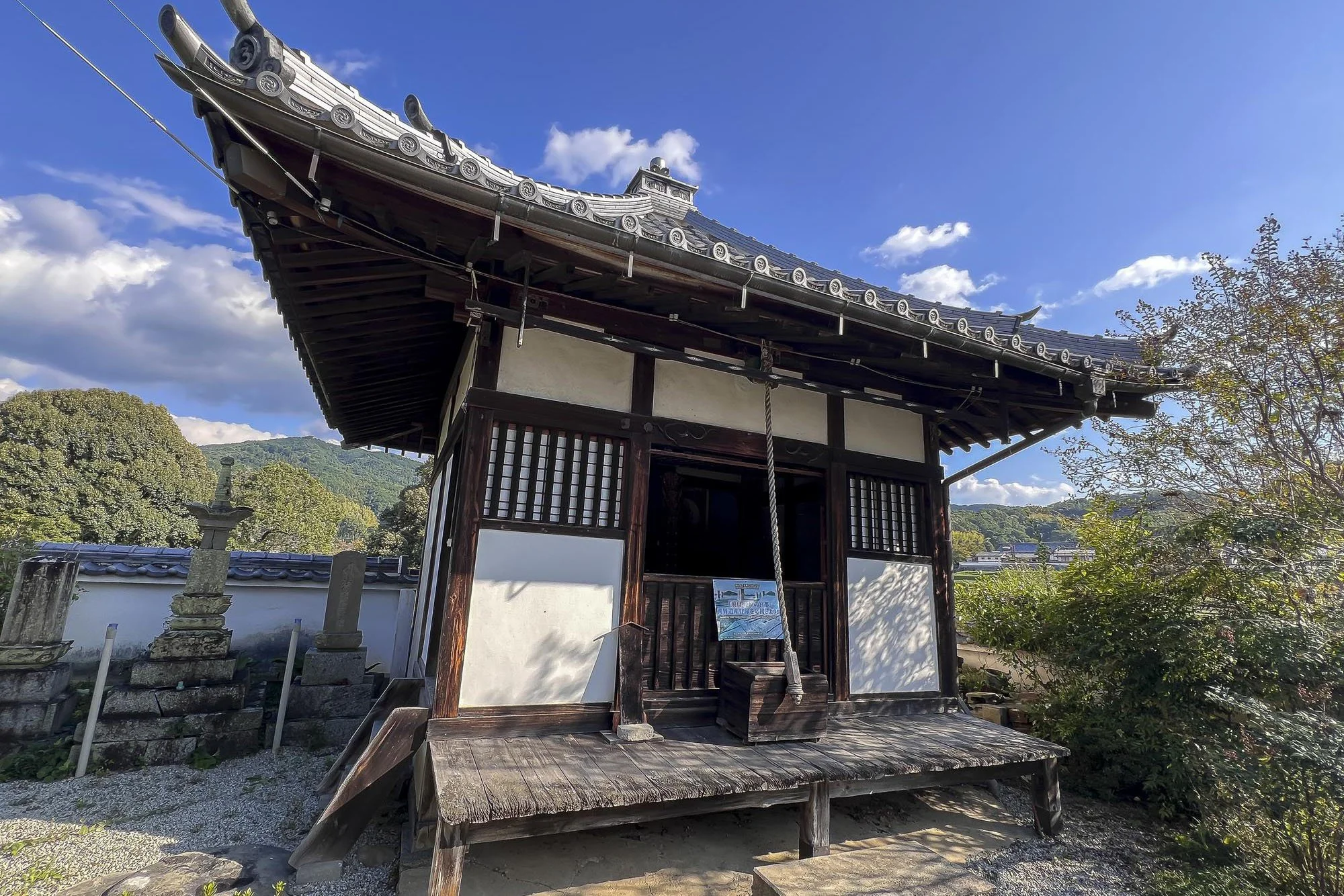 A traditional Japanese wooden temple with a tiled roof, situated outdoors with trees and a mountain in the background under a partly cloudy sky.