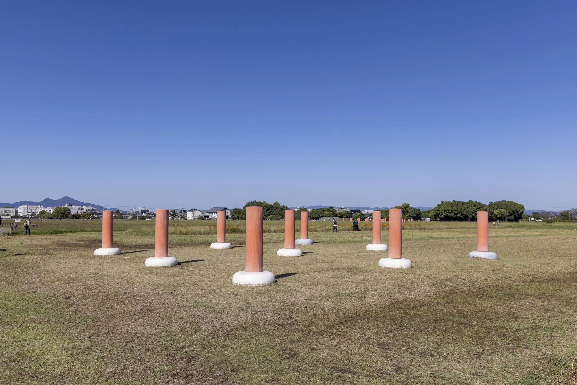 Open grassy field with nine tall, pinkish-orange cylindrical pillars on round white bases, under a clear blue sky, with a few distant trees and buildings in the background.