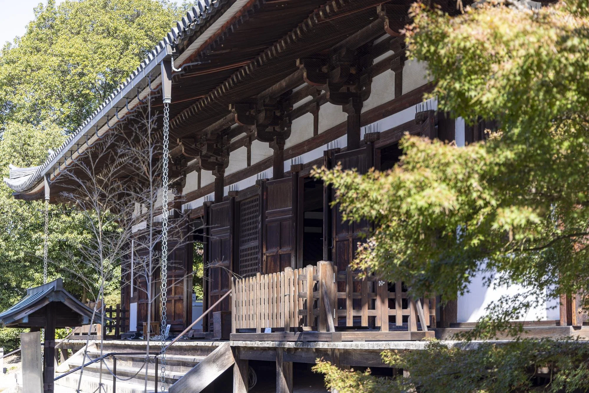 Traditional Japanese wooden temple with open sliding doors, surrounded by green trees and a small wooden fence, set against a blue sky.