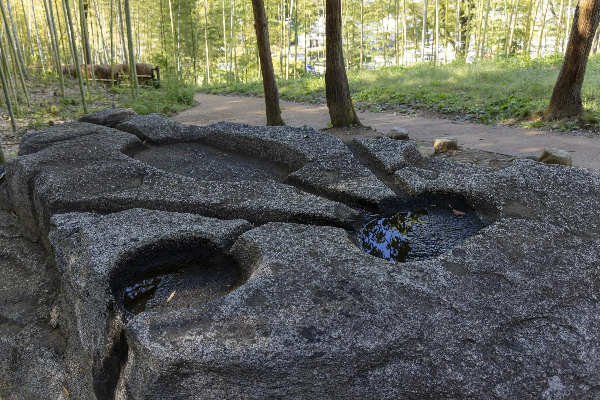 A stone water basin with two small pools of water, located in a wooded area with a dirt path and trees in the background.
