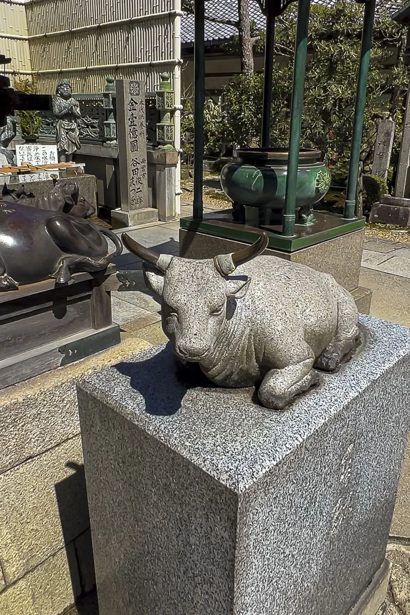 A stone sculpture of a reclining bull with curved horns, situated on a granite pedestal in a traditional Japanese setting, with other statues, a stone monument, and a green and black decorative urn in the background.