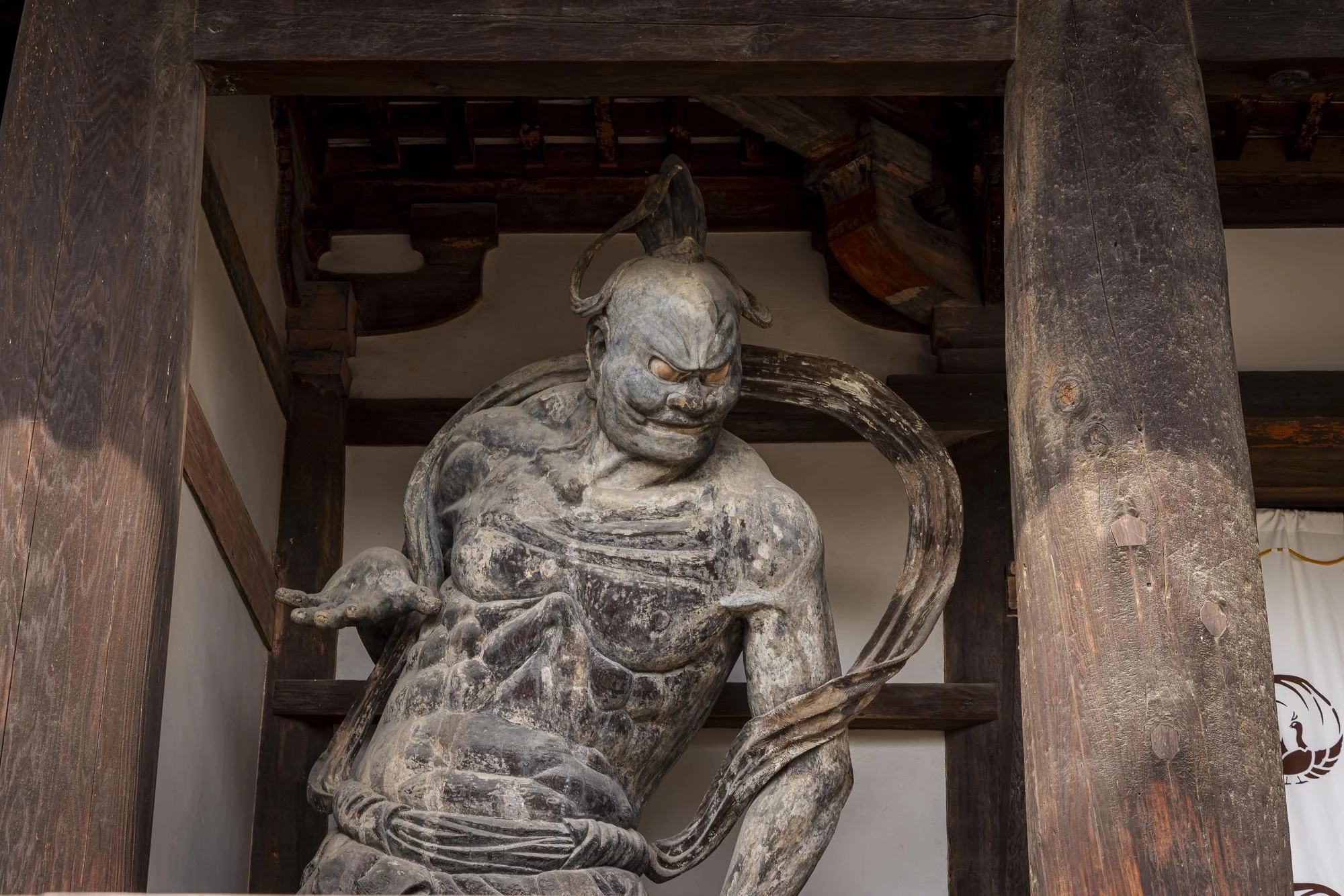 A weathered wooden sculpture of a fierce warrior or deity with an intense expression, pointing with one hand and holding a curved weapon in the other, surrounded by large wooden pillars in a traditional setting.