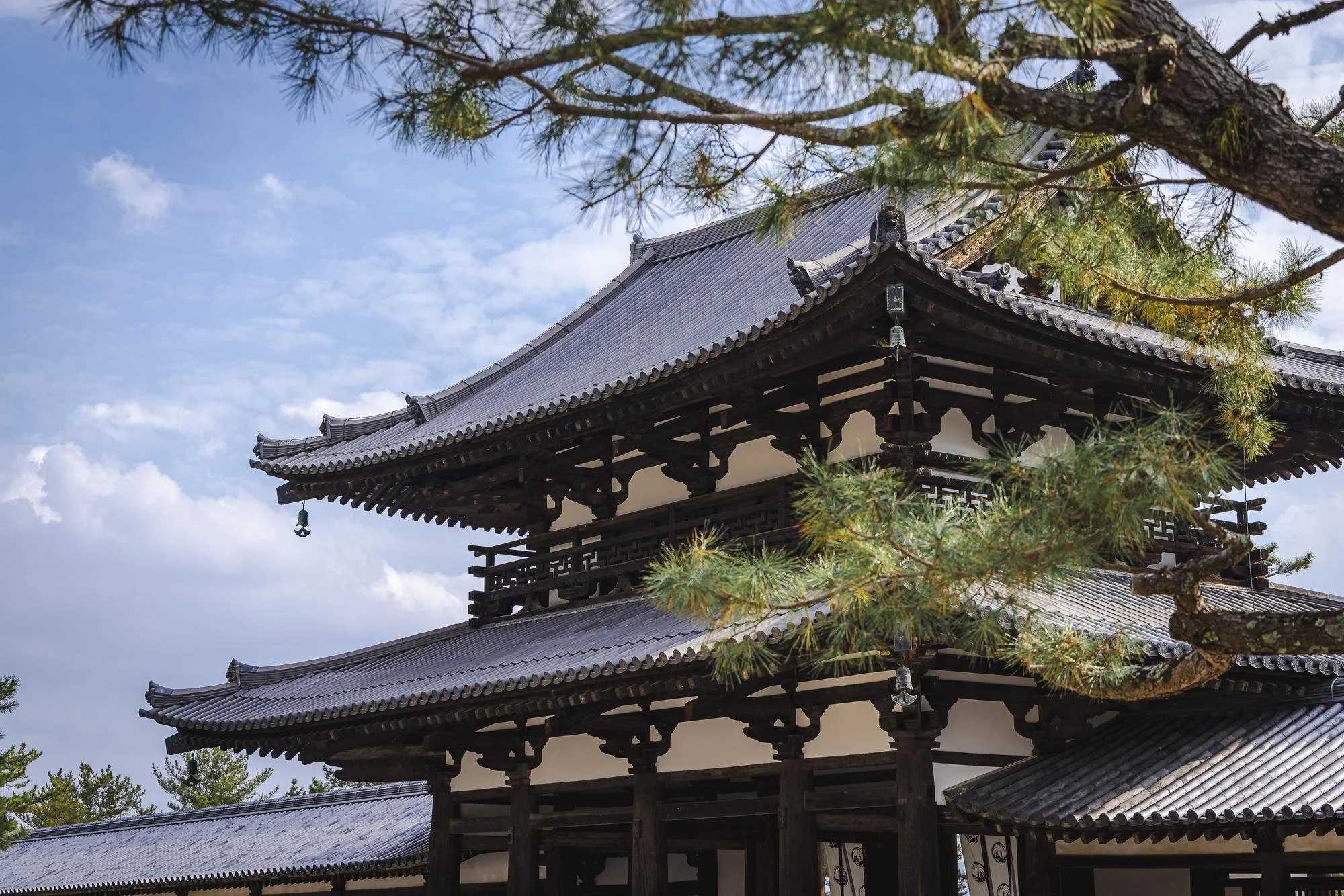 A traditional Japanese temple with a curved tiled roof, surrounded by green pine trees, under a partly cloudy blue sky.