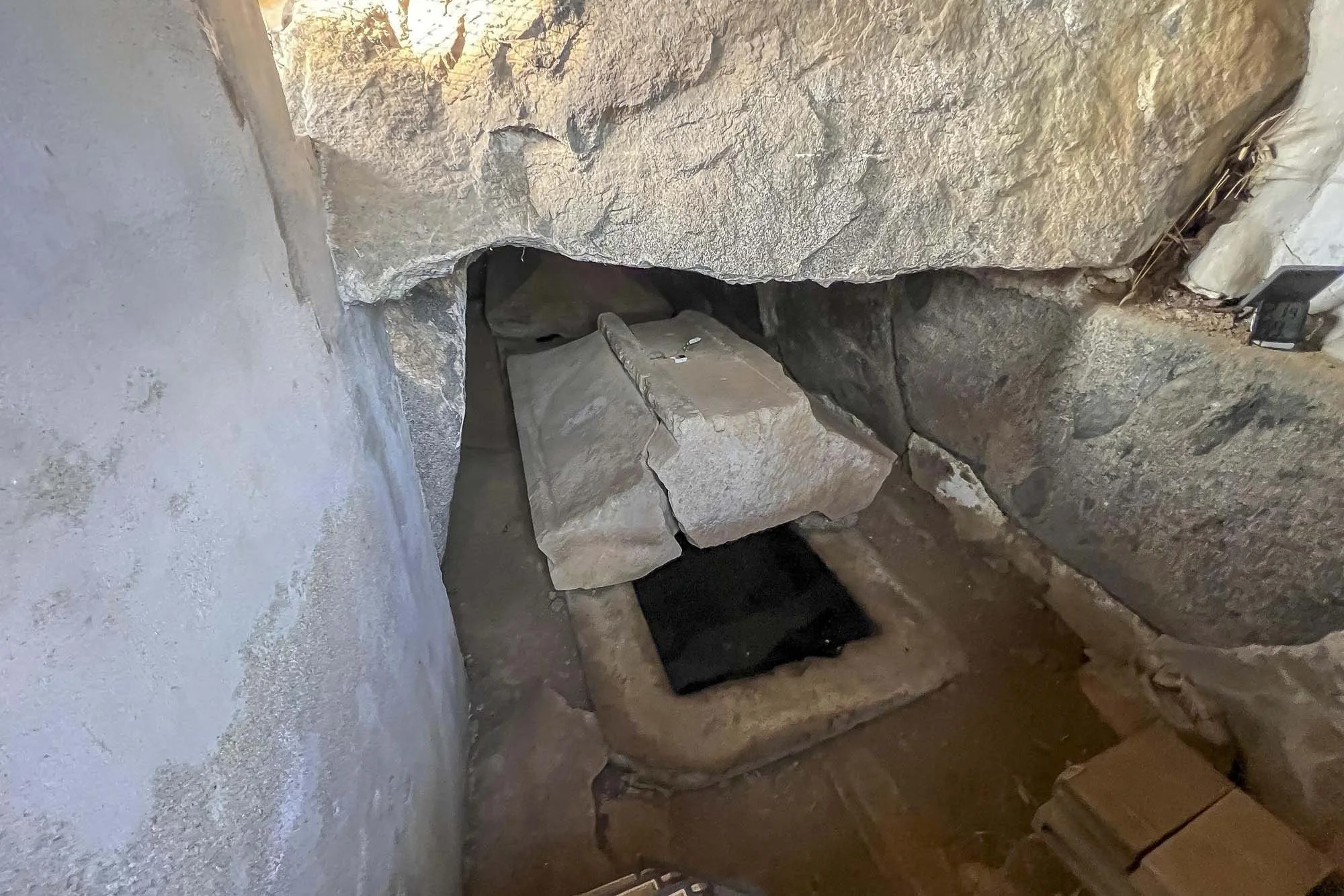 Shobuike Mounded Tomb: A small stone altar or shrine built into a stone wall, with a rectangular stone block on top, and a black stone or metal object beneath, set in a corner with walls made of large rocks and concrete.