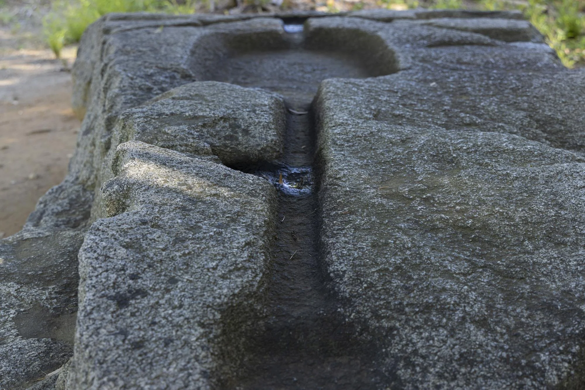 Close-up of a stone water fountain with a carved channel and a small stream of water flowing through it, outdoors with blurred greenery in the background.