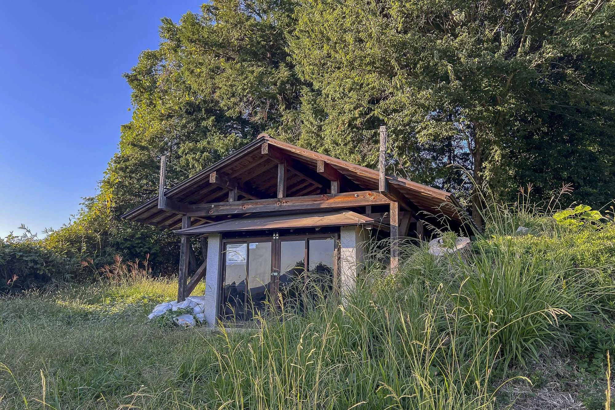 Small wooden house in a grassy area with tall grass and large trees in the background.