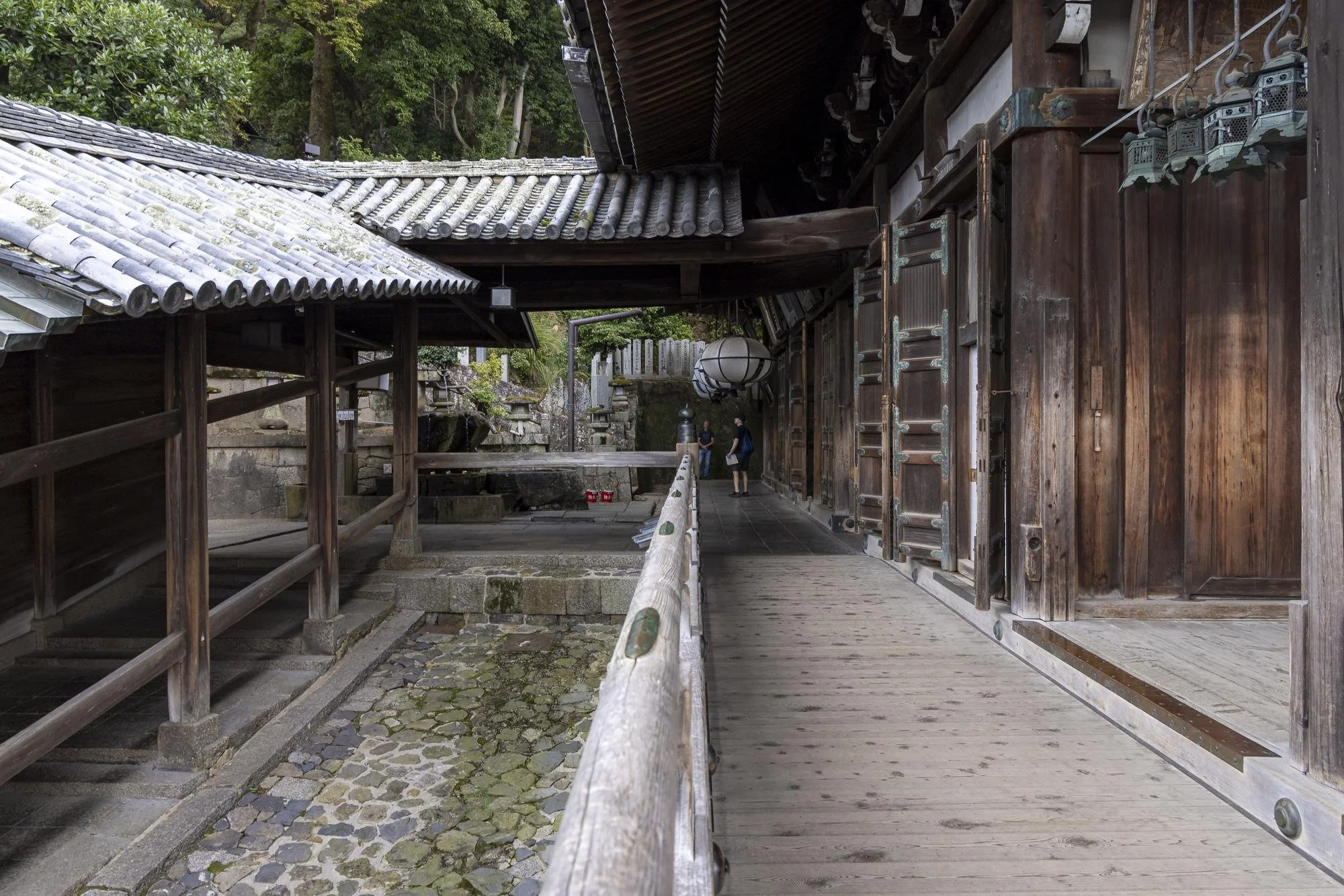 Traditional Japanese temple with wooden architecture, sliding doors, lanterns, and stone lanterns in the background, surrounded by greenery.