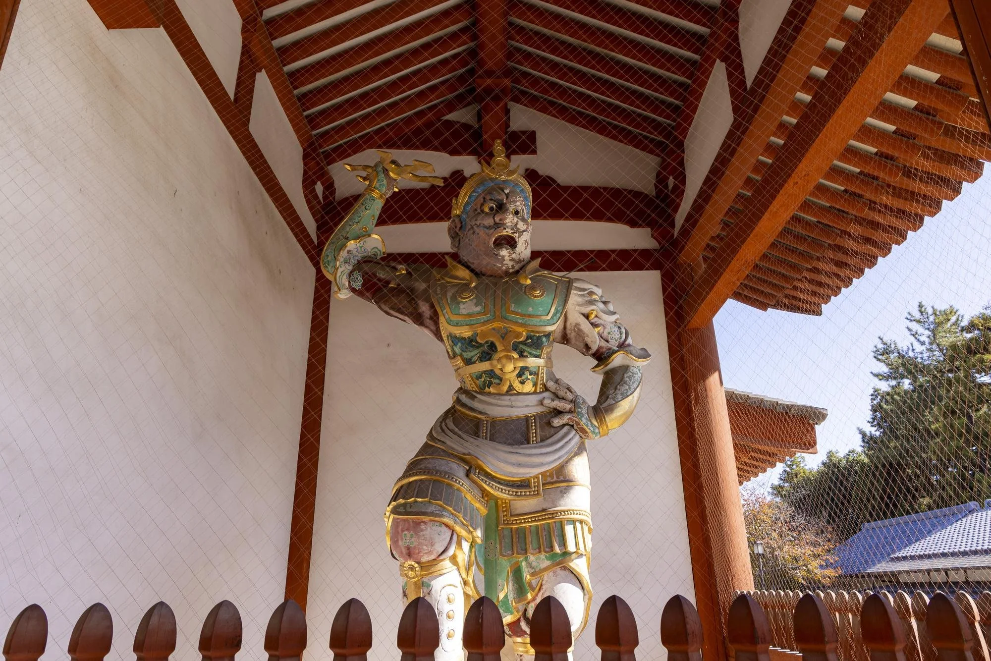 Colorful statue of a fierce figure, possibly a mythological or religious character, inside a traditional wooden structure with a roof, surrounded by a wooden fence, and trees outside.