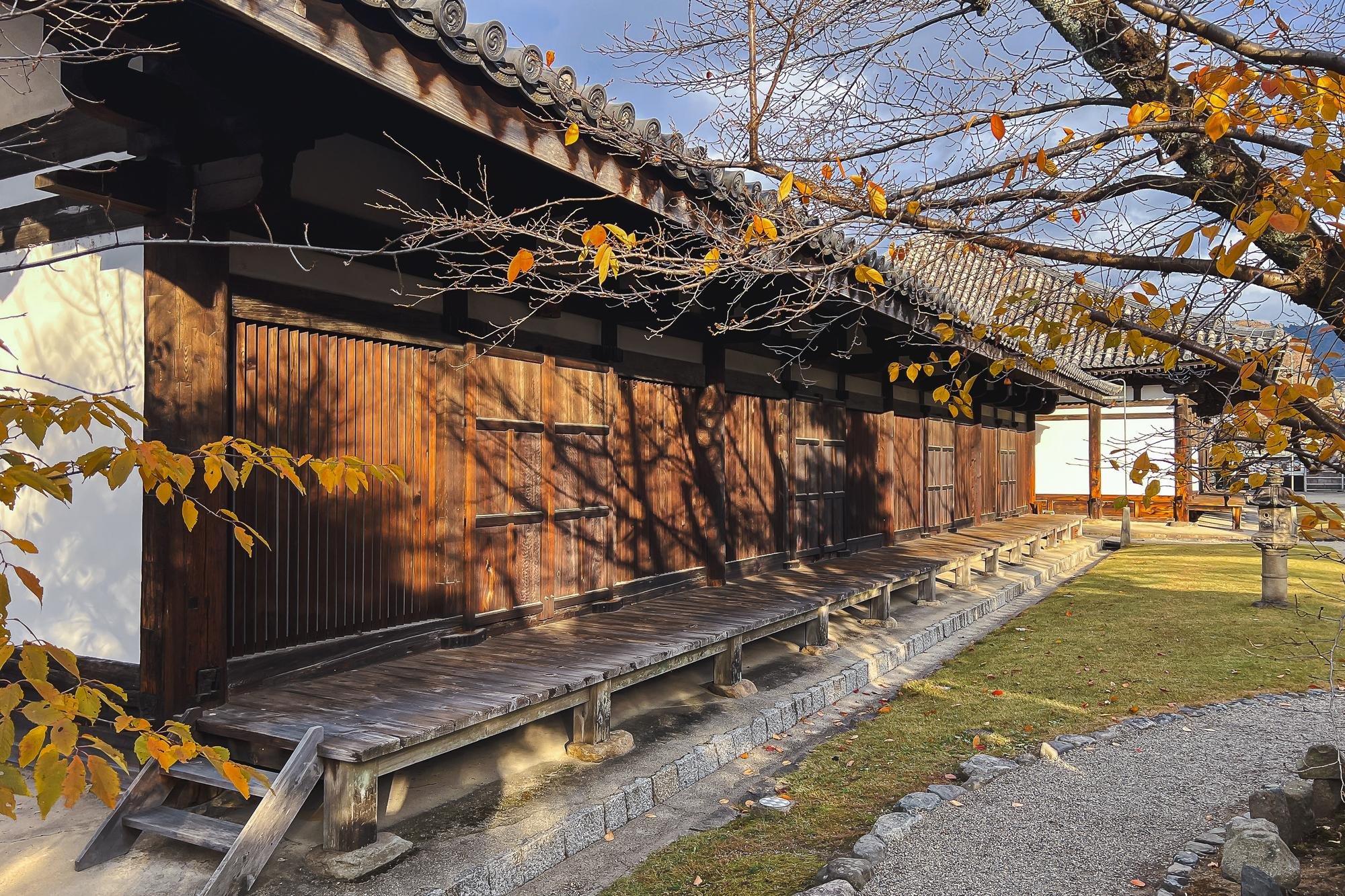 Traditional Japanese wooden building with a tiled roof, surrounded by autumn trees with orange and yellow leaves, and a stone lantern on the right.