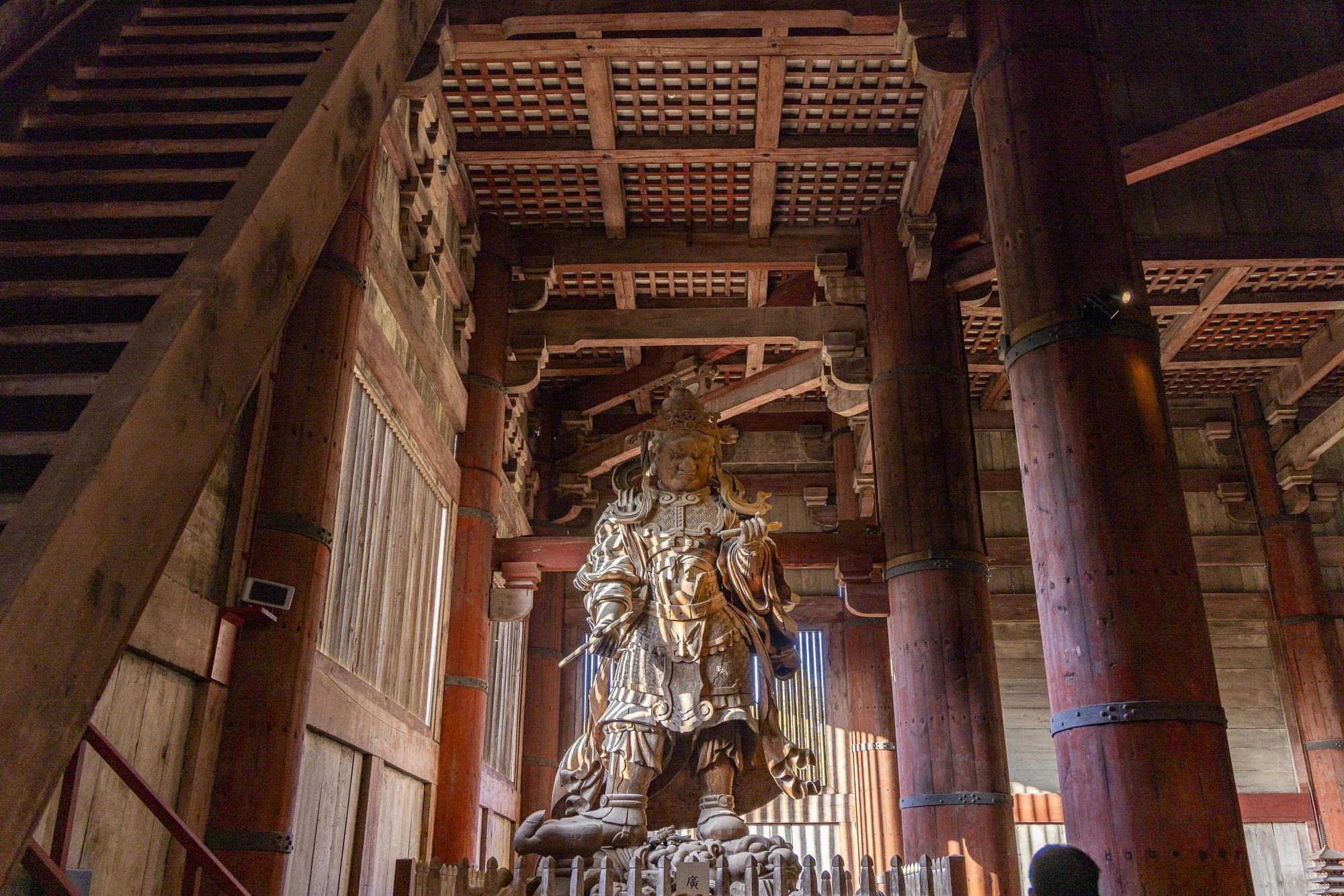 Inside a traditional wooden temple, a large wooden statue of a fierce warrior deity stands on a platform, surrounded by tall wooden pillars and beams.