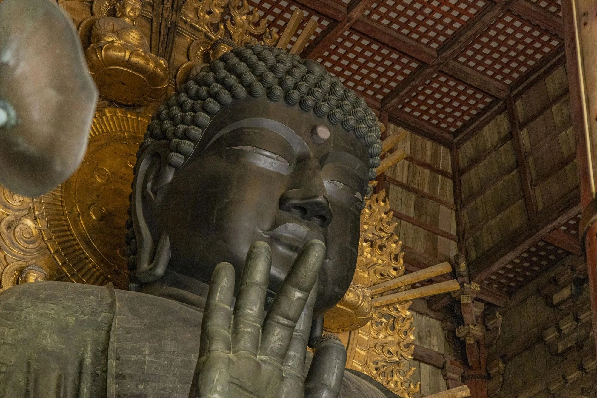 Close-up of a large bronze Buddha statue inside a wooden temple, with the Buddha's right hand raised in a gesture of blessing and the left hand resting on the lap, intricate gold decorations behind.