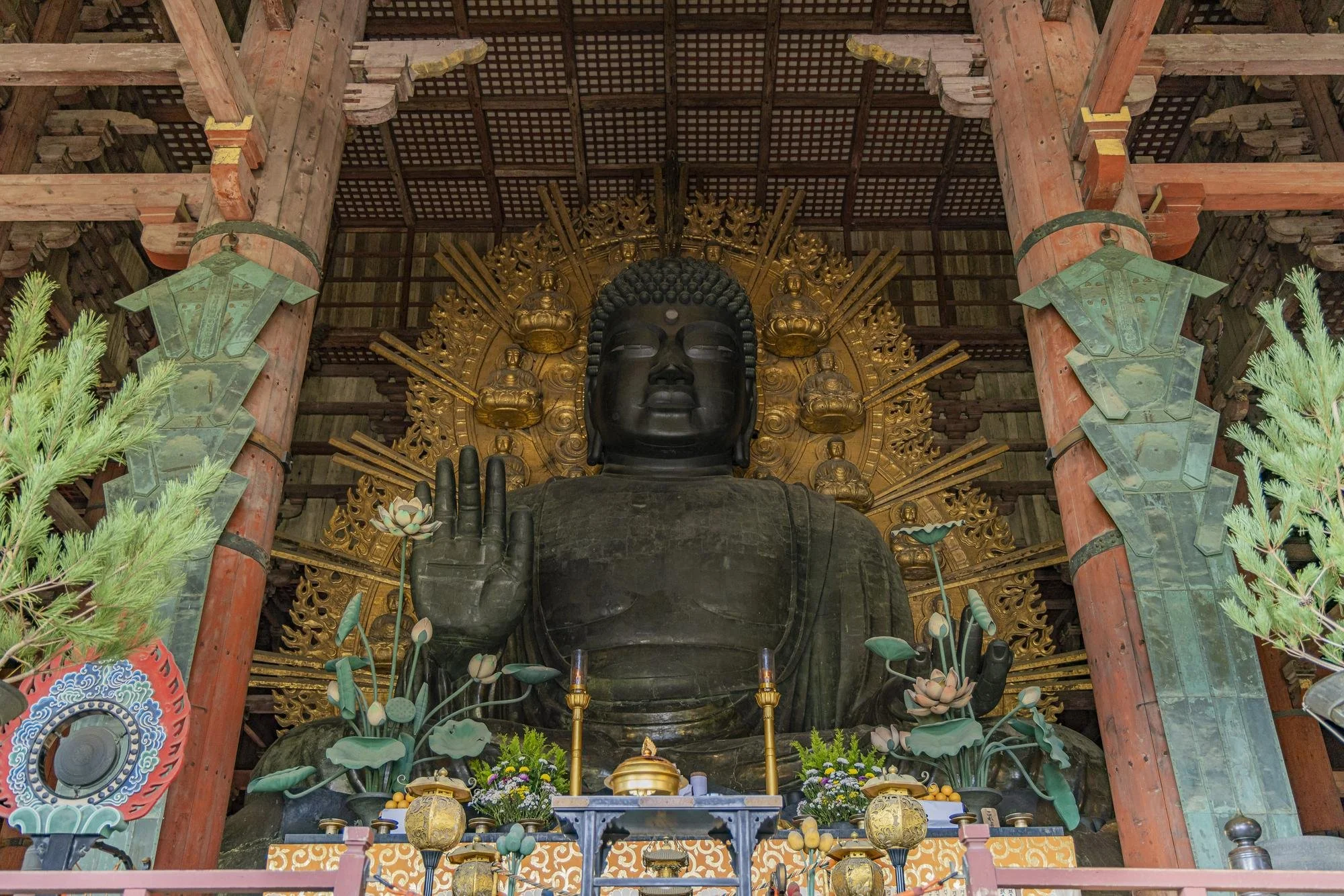 Large black Buddha statue inside a wooden temple, with a multi-layered gold background, surrounded by plants and decorative items on an altar.