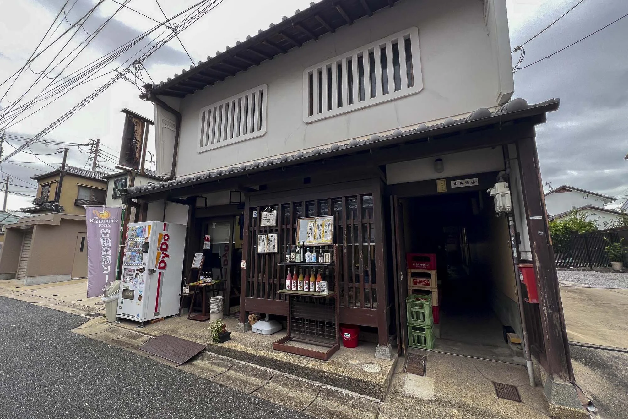 Traditional Japanese building with wooden lattice and jars outside, vending machine nearby, street with power lines overhead, cloudy sky.
