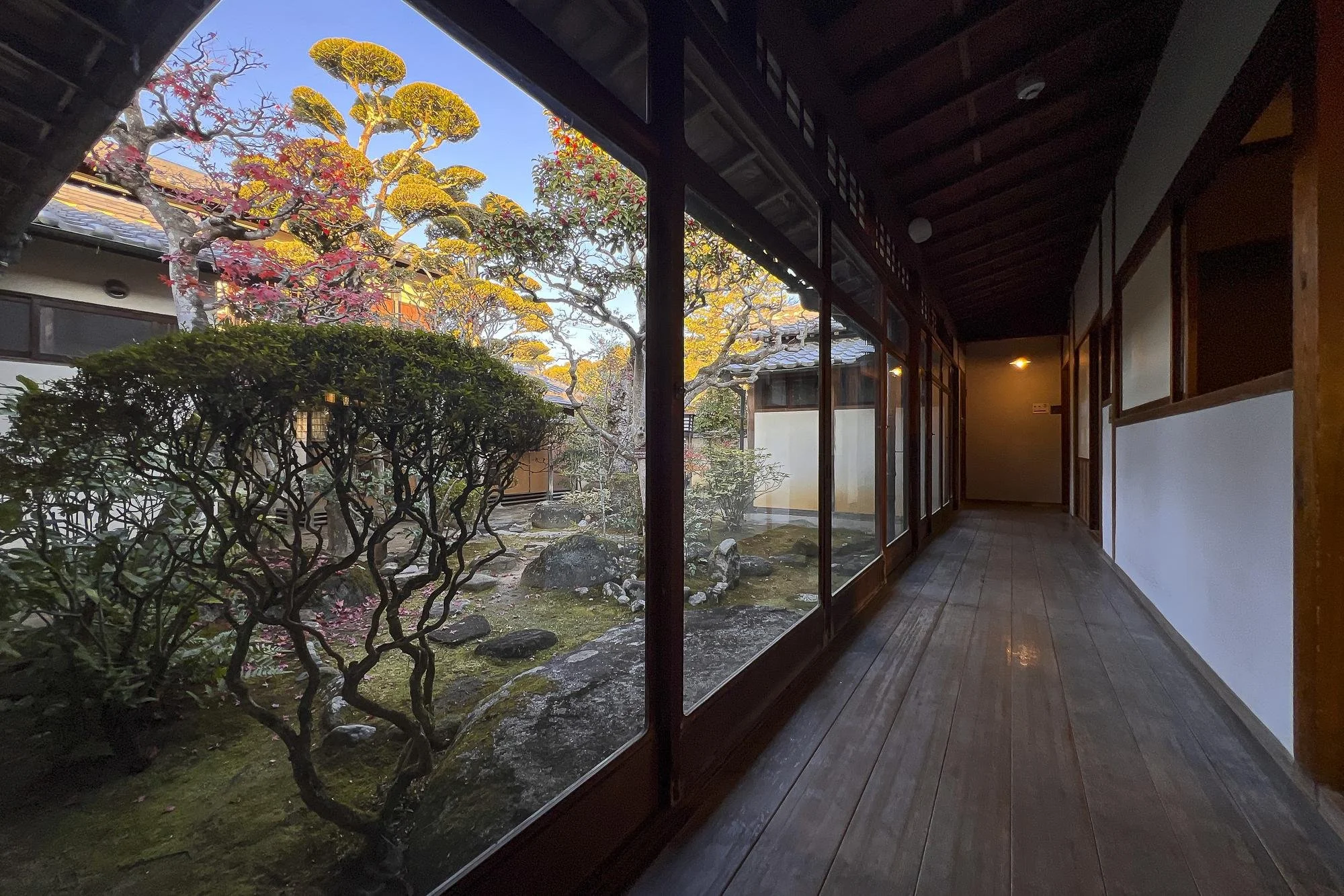 View of a traditional Japanese garden with trees and rocks, seen through large glass windows from a wooden corridor with sliding doors and white walls.