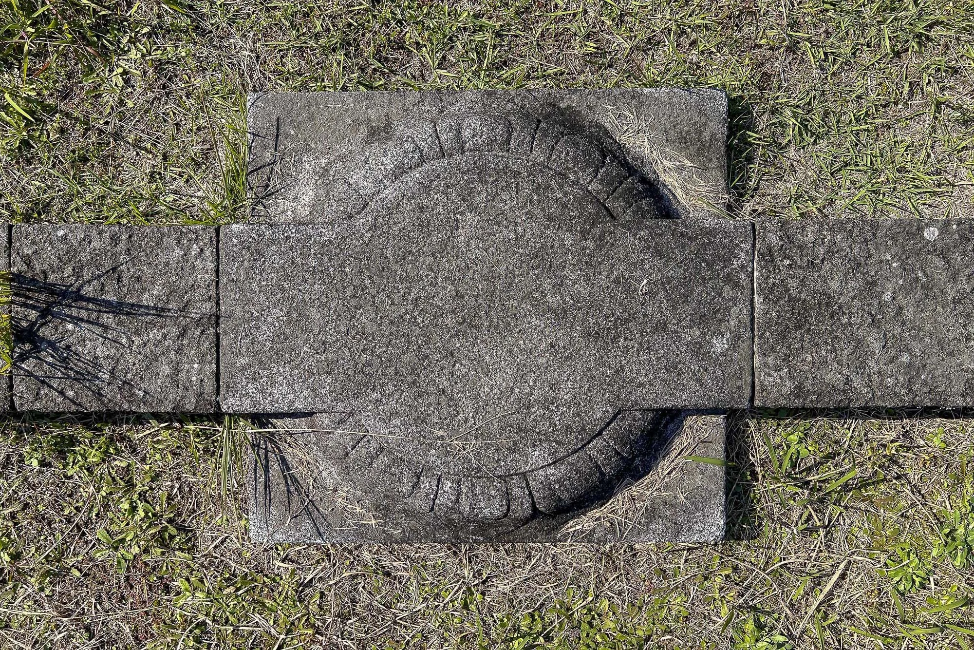 A stone or concrete circular form with an arrow carved into it, surrounded by three rectangular stones, on grass with small plants.