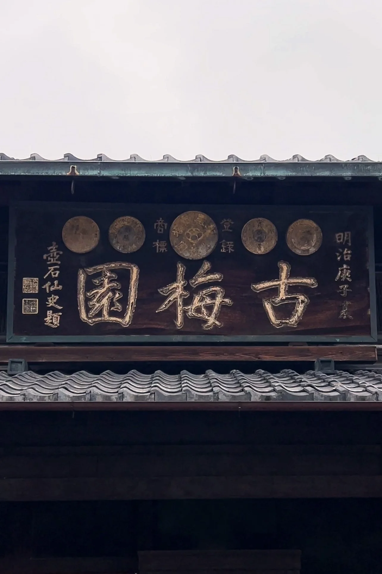 A signboard with Japanese characters and circular gold medals or coins mounted on a wooden structure under a tiled roof.