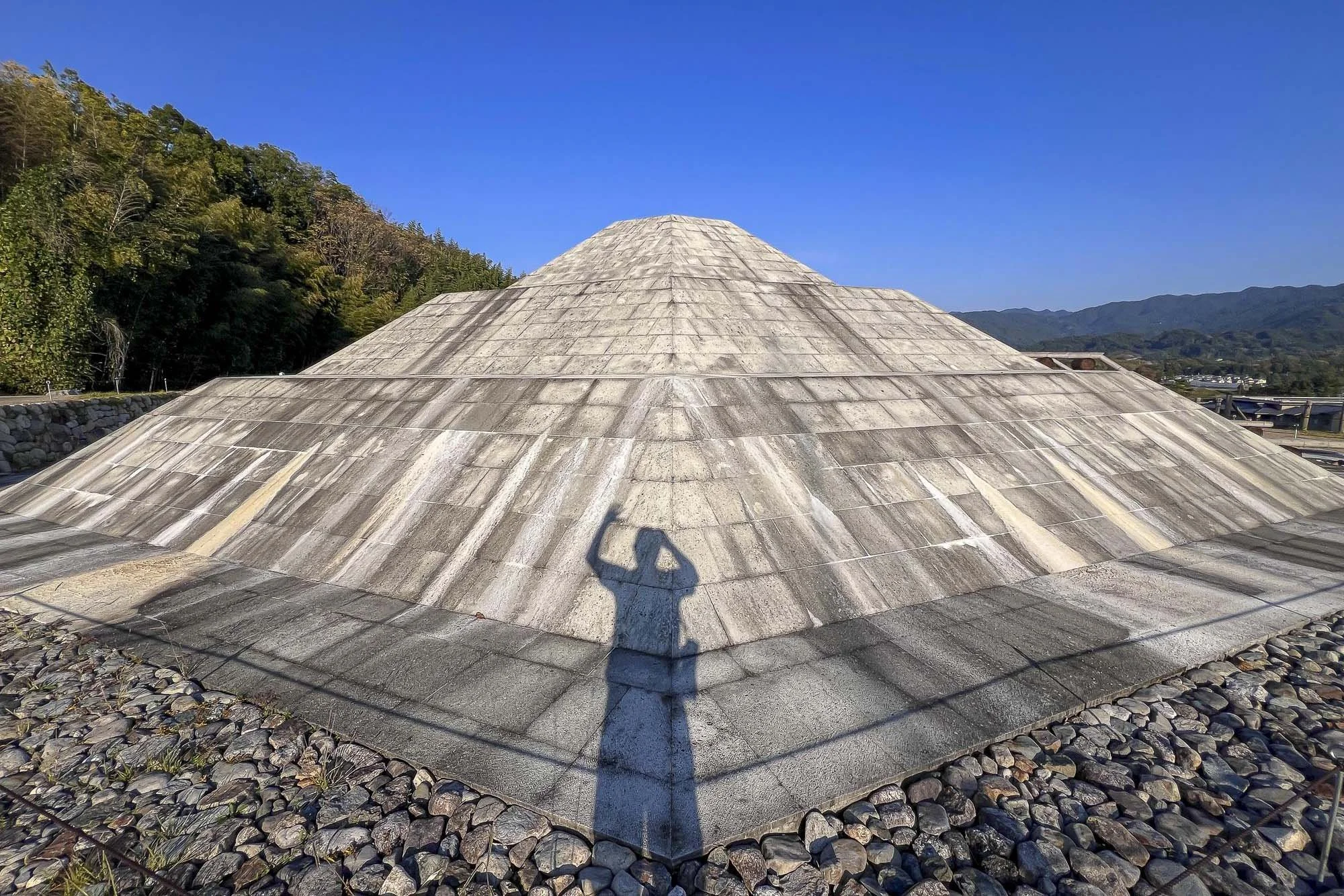 Photo of a large, multi-tiered, stone-tiled roof with a mountain and blue sky in the background. The shadow of a person taking a photo is visible on the roof.