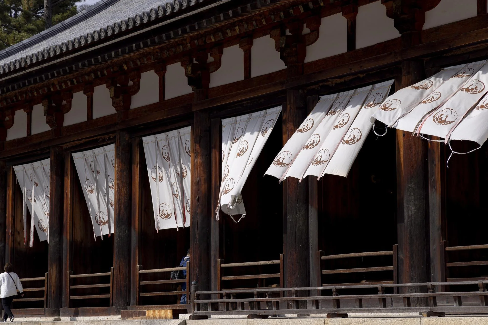 Traditional Japanese temple with wooden architecture and white curtains with brown emblem designs hanging in openings, a person in white walking nearby.