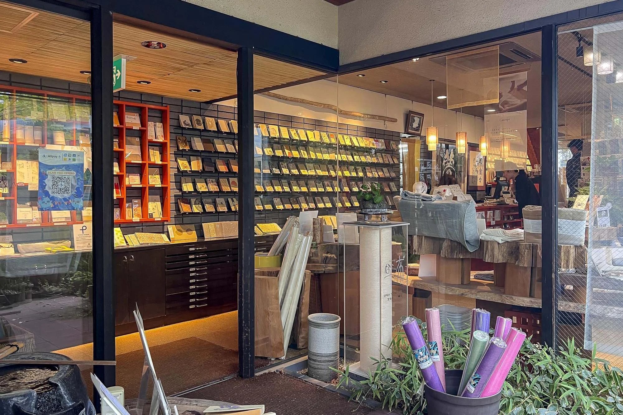 View of a bookstore or shop through a glass window, showing shelves of books and magazines, a counter with a person attending, and decorative items inside.
