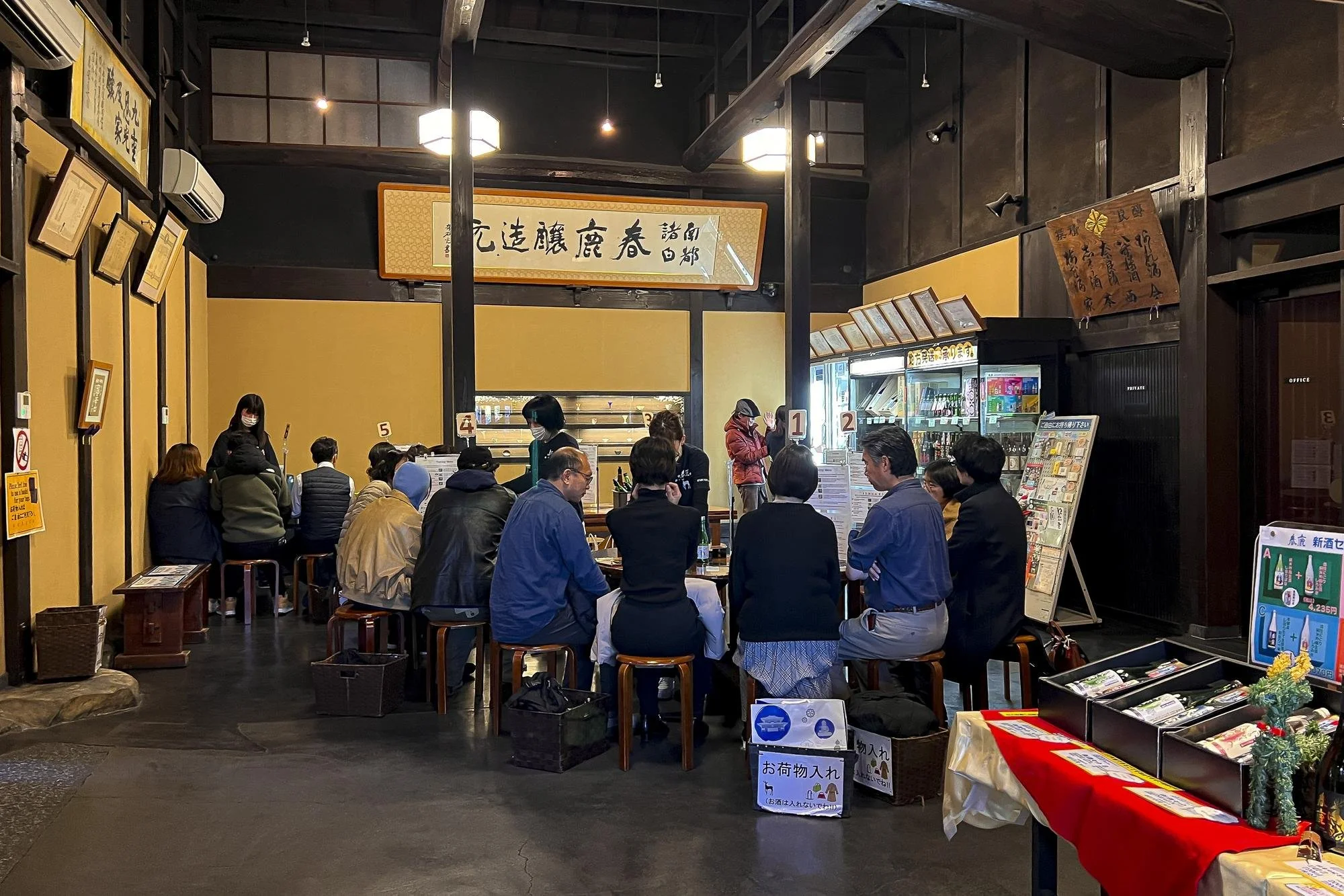 Traditional Japanese building with white walls and wooden accents, featuring a small entrance, a display window, and decorative elements including a paper lantern and a large sake barrel outside.