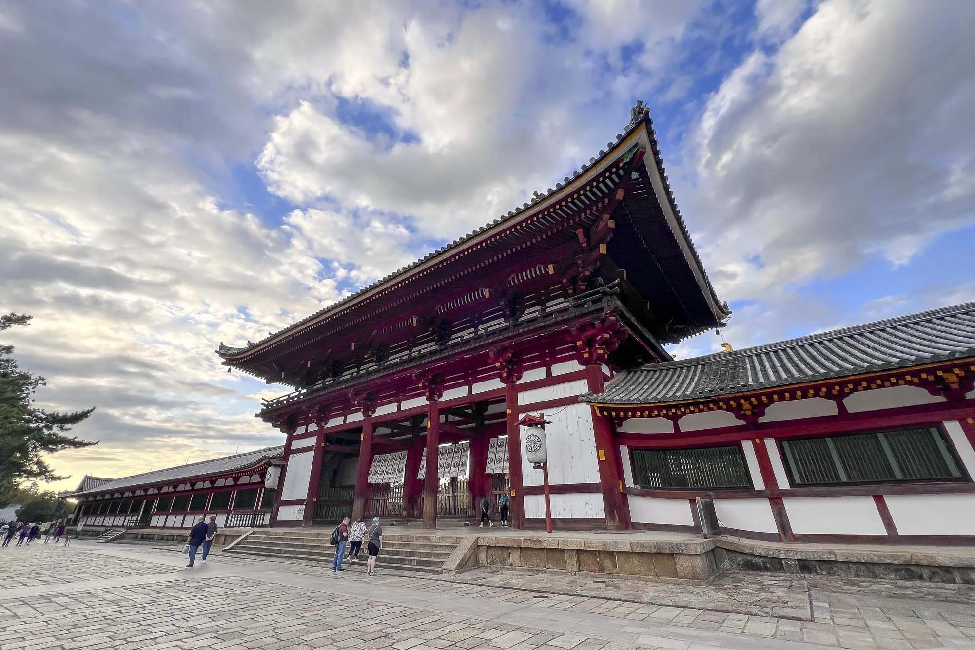 A traditional Japanese temple with a large entrance gate featuring red pillars and a multi-tiered roof, people walking around the temple grounds, under a partly cloudy sky.