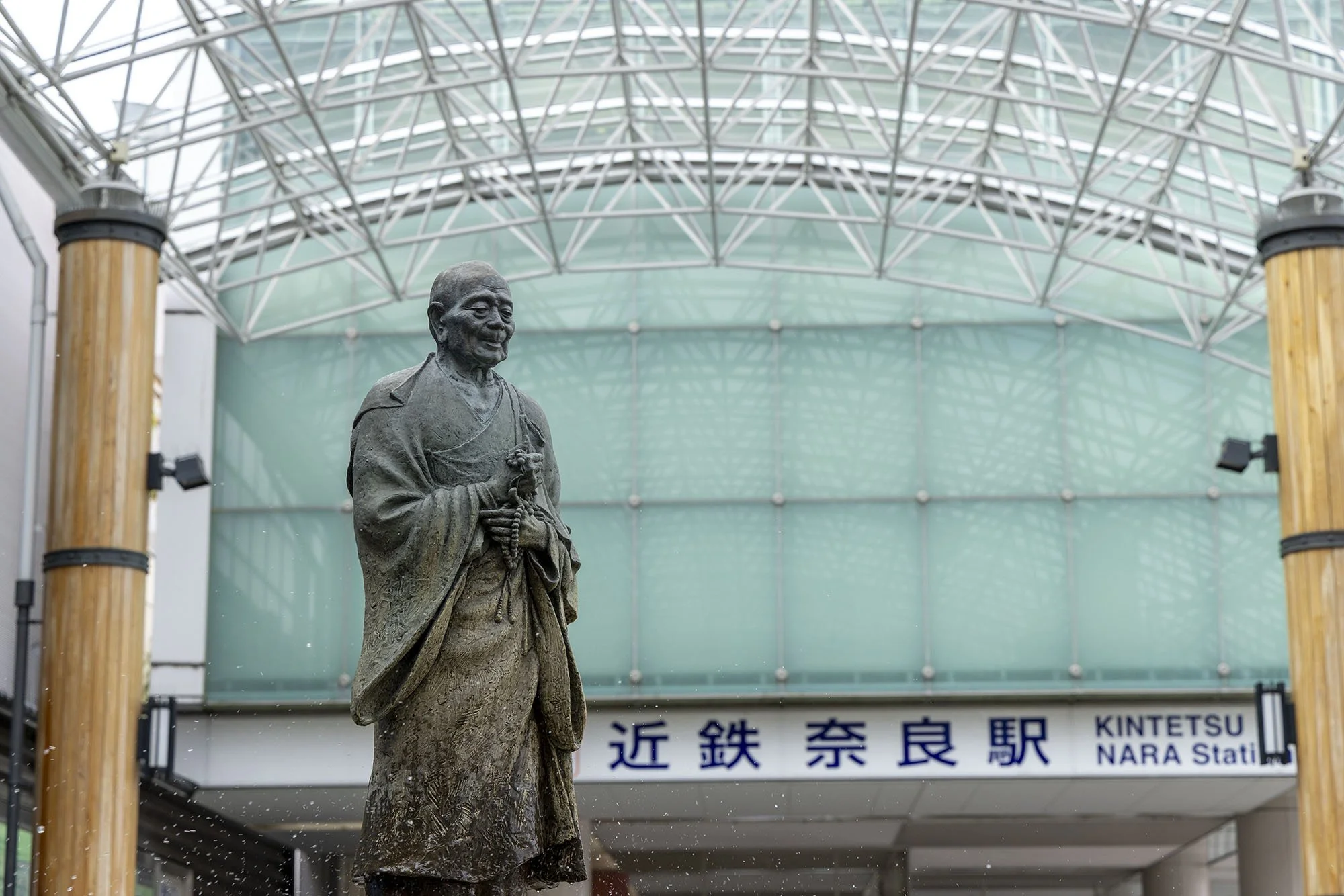 Bronze statue of Gyōki standing beneath the glass canopy outside Kintetsu Nara Station, with the station sign visible in the background.