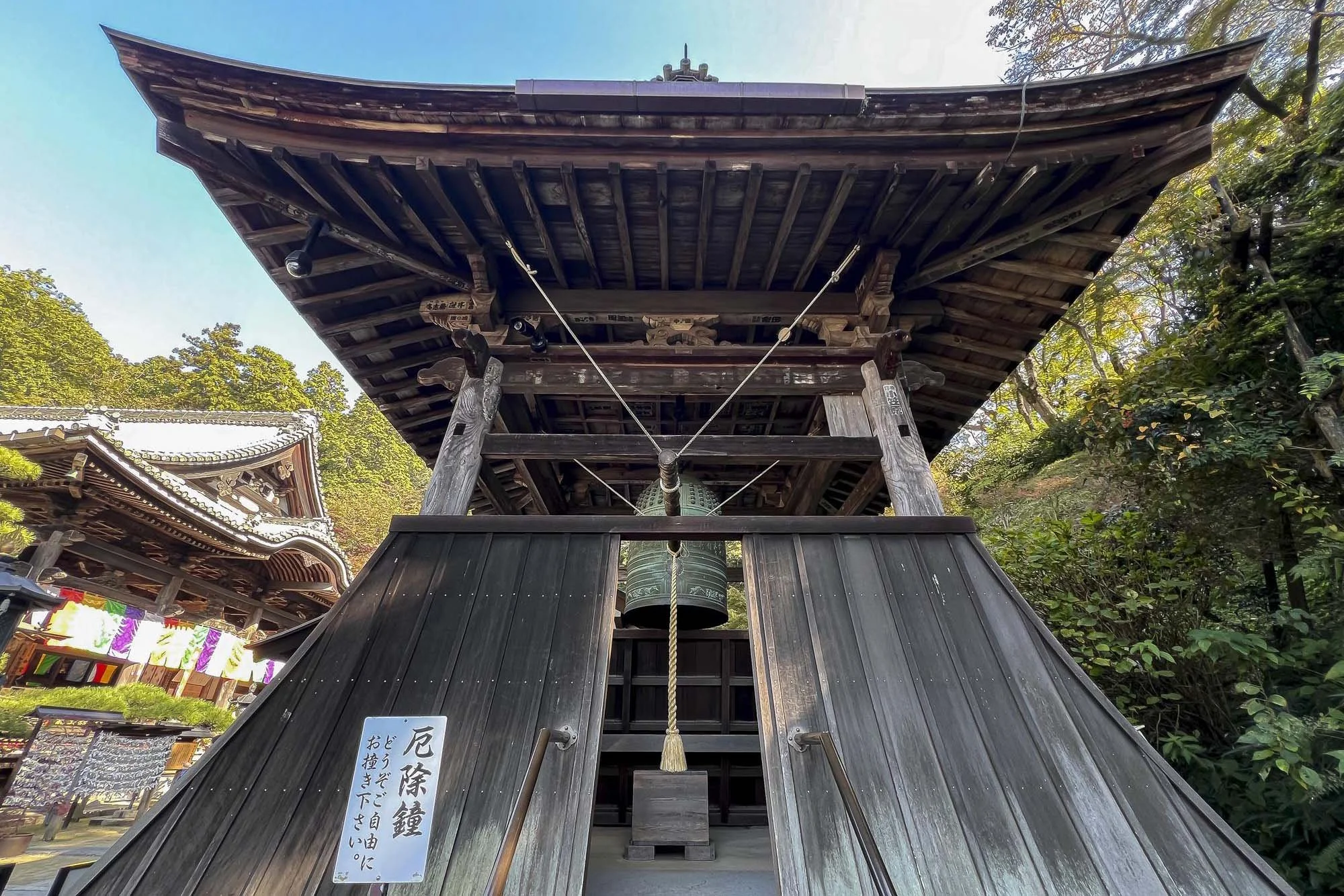 A traditional Japanese temple bell hanging inside a wooden structure with a large rope hanging down in front, surrounded by trees and other temple buildings.