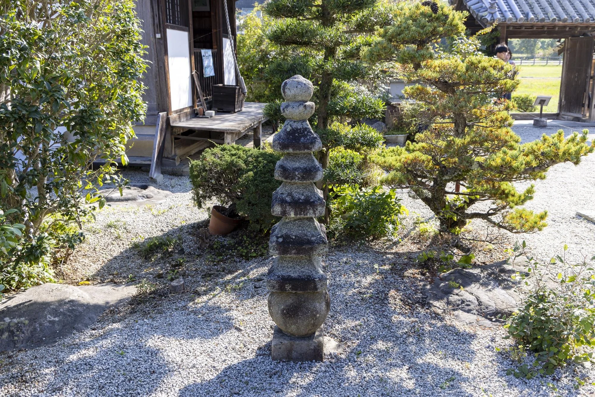 A traditional Japanese garden featuring a stone pagoda sculpture at the center, surrounded by manicured greenery and trees. There is a wooden building with a porch in the background, and a person is visible in the distance. The ground is covered with