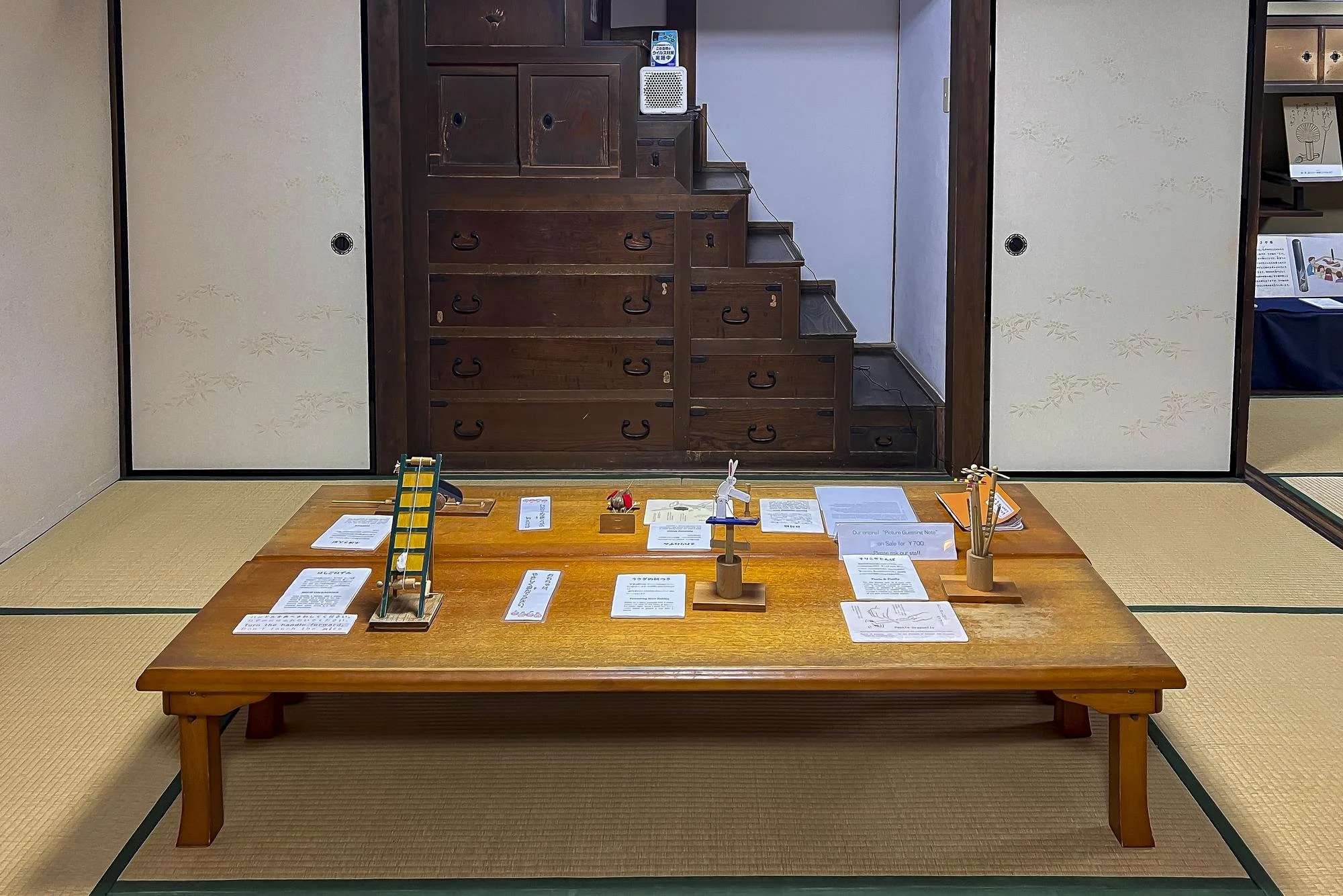 A traditional Japanese room with tatami mats, a low wooden table with various papers and decorative items, sliding doors with floral patterns, and a staircase in the background leading to a second level.