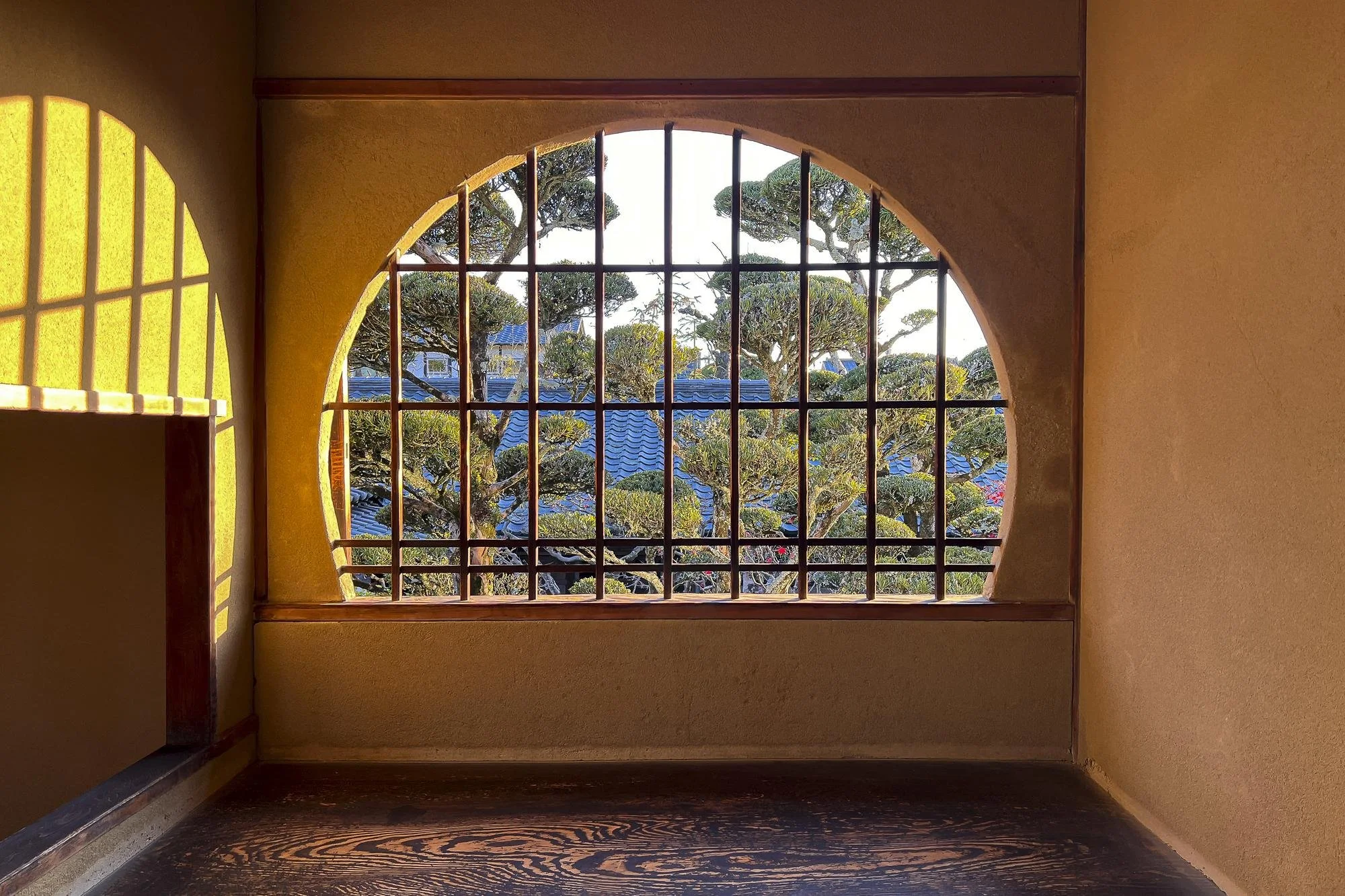 Interior of a room with a large arched window with grid bars, showing trees and blue rooftops outside, with warm sunlight casting shadows inside.