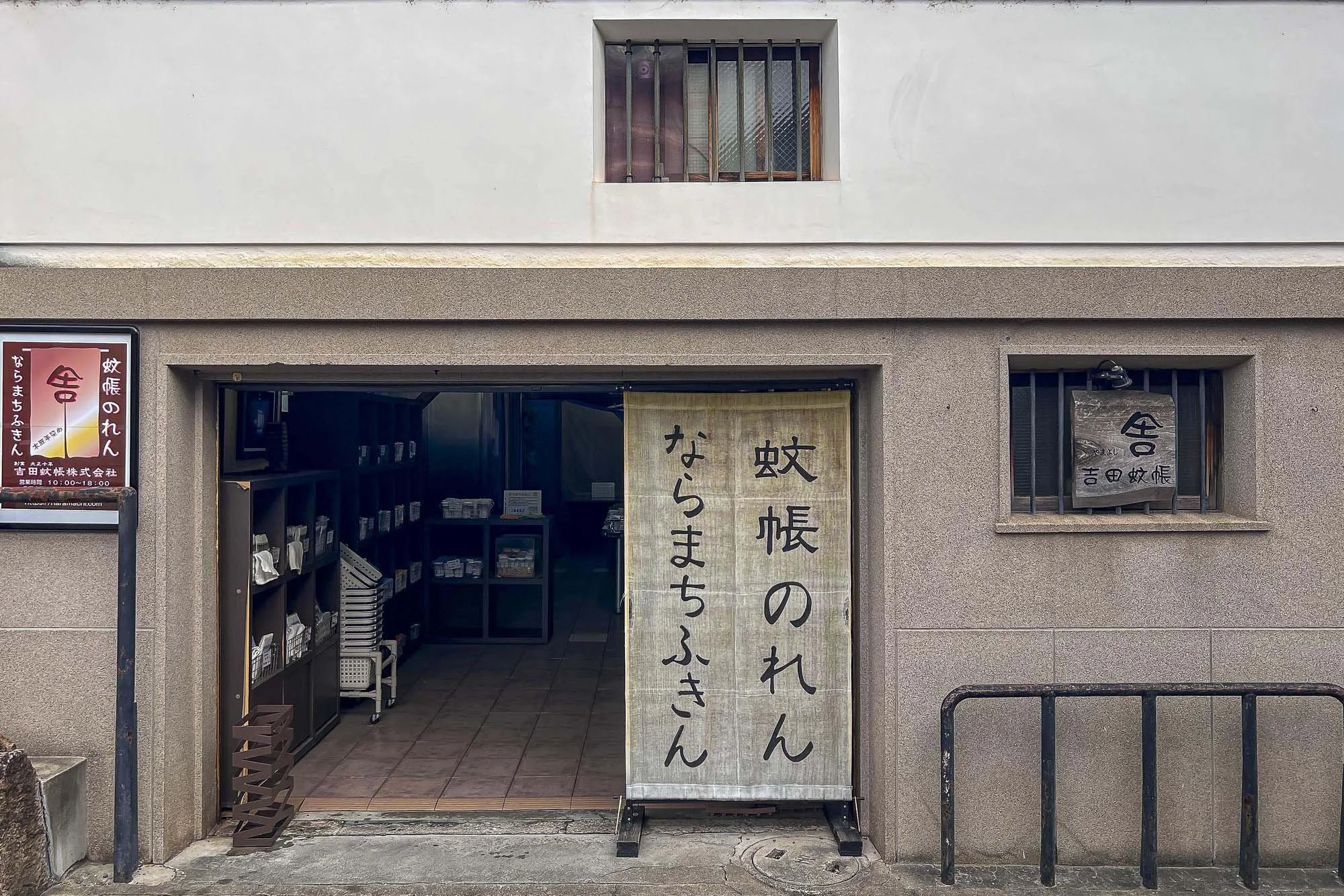 The entrance of a Japanese store with a beige cloth curtain hanging over the sliding door. The curtain has black Japanese characters on it. To the left, there are shelves with small packages, and to the right, a small window with a dark-colored sign 