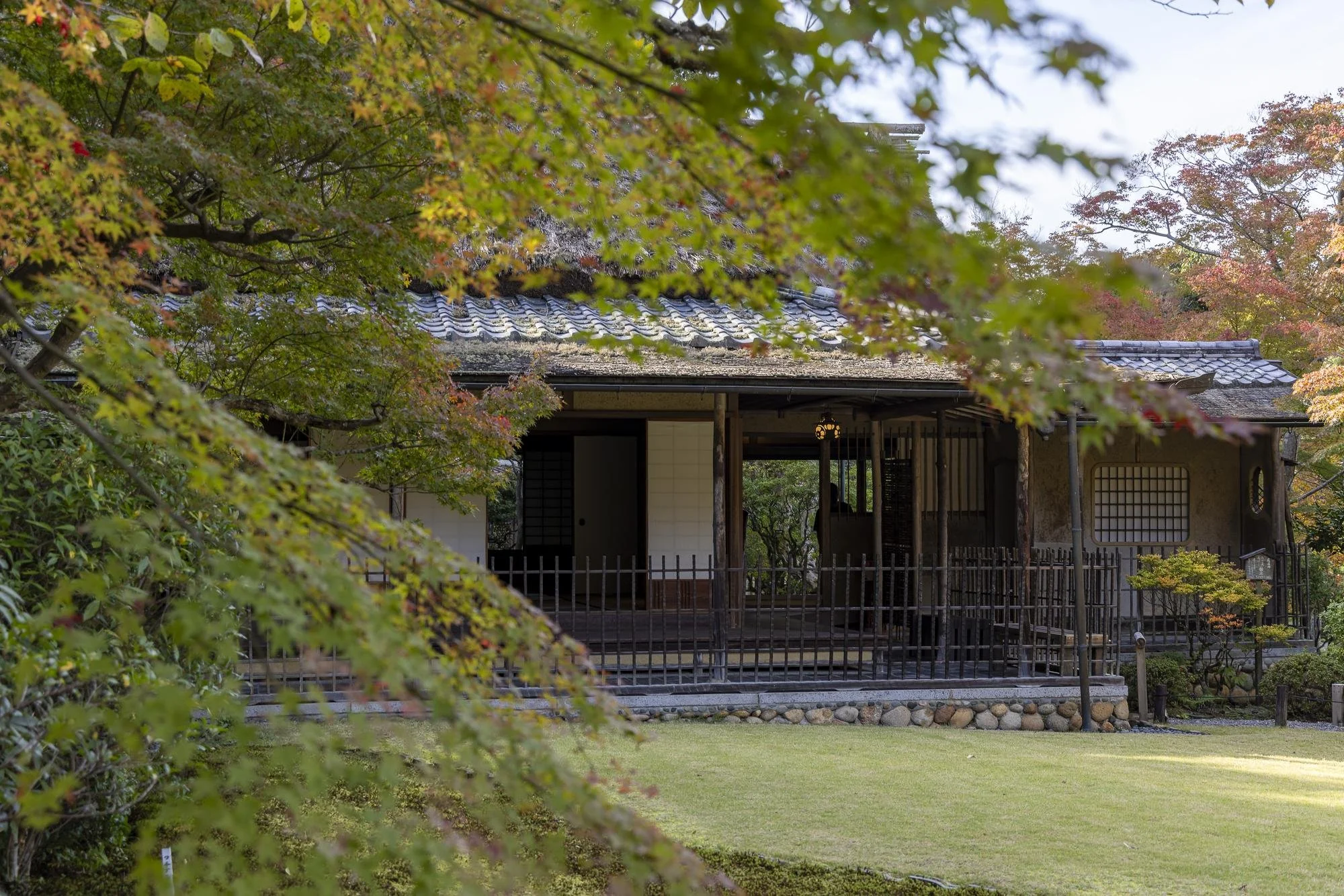 Traditional Japanese house with wooden structure, sliding doors, and tiled roof, surrounded by autumn foliage and green grass