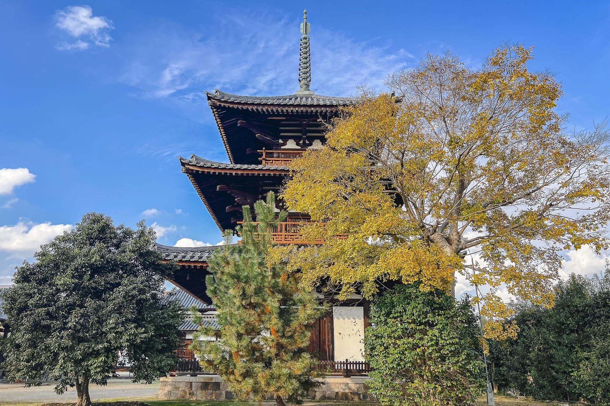 Traditional Japanese pagoda with multiple tiers, surrounded by trees with yellow and green leaves, under a blue sky with clouds.