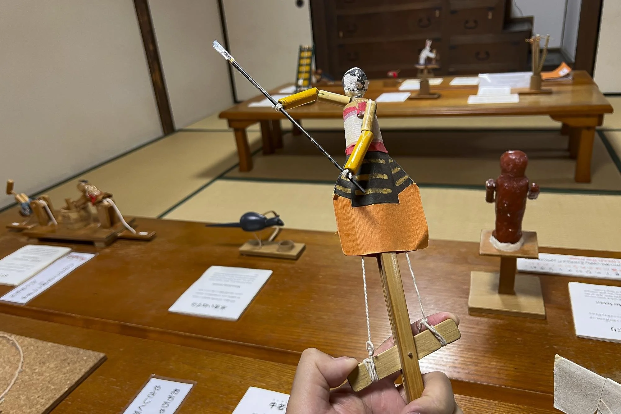 A person's hand holding a miniature wooden doll with a painted face, wearing an orange and black skirt and holding a spear, displayed in a museum with other small wooden figures on tables.