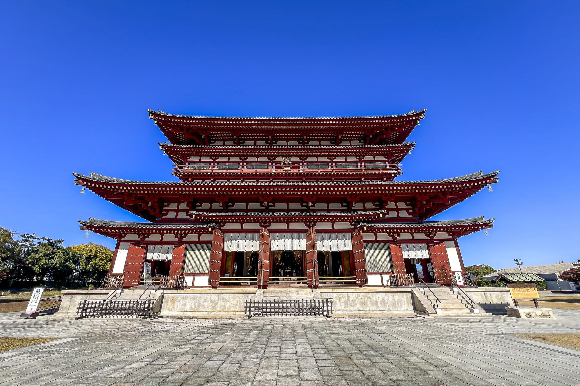 A traditional Japanese temple with multiple tiered, curved roofs, red and white walls, and wooden details, set against a clear blue sky.