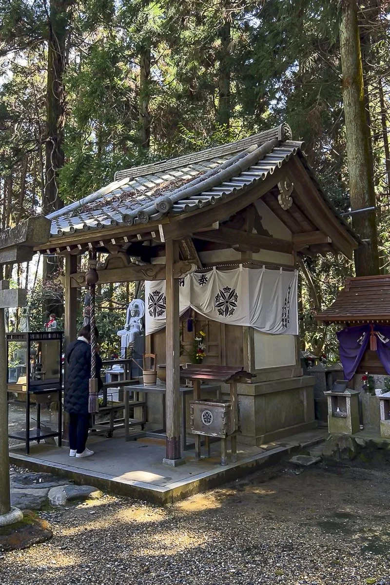 A small traditional Japanese shrine with a wooden structure, tiled roof, and white curtains decorated with black symbols, surrounded by trees and foliage.