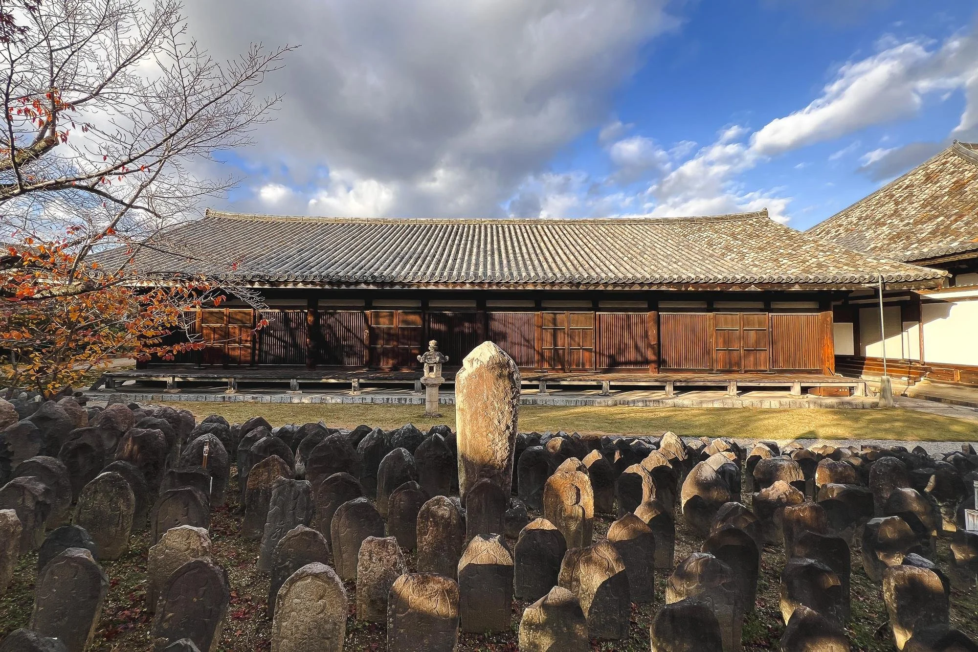 A traditional Japanese building with a tiled roof and wooden walls, surrounded by a garden with stone statues and a small lantern, under a partly cloudy sky.