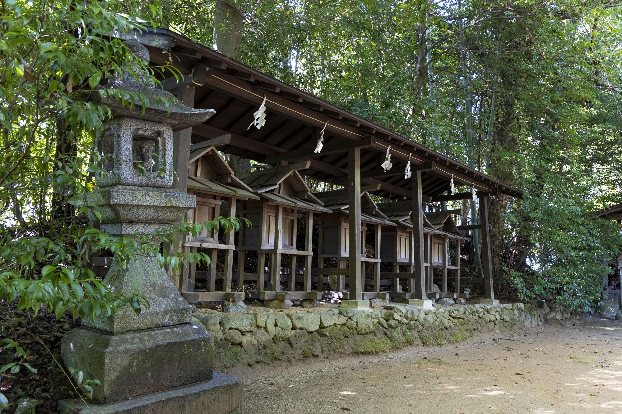 A series of small wooden shrine structures with roofs, lined up on a stone foundation in a forested area, with a traditional stone lantern in the foreground.