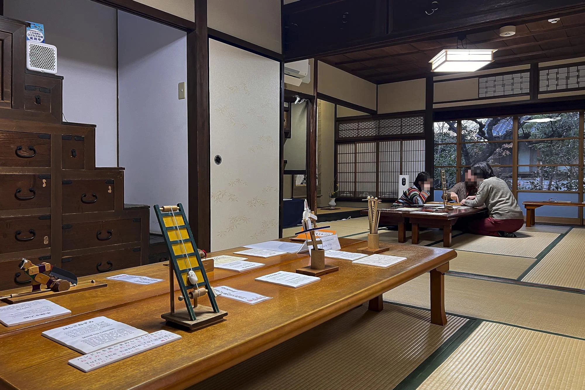 Traditional Japanese room with tatami mats, low wooden tables, and sliding shoji doors. Two people are seated on the floor at one table, engaging in an activity. There are papers on the tables, and a decorative display with wooden figures is visible.