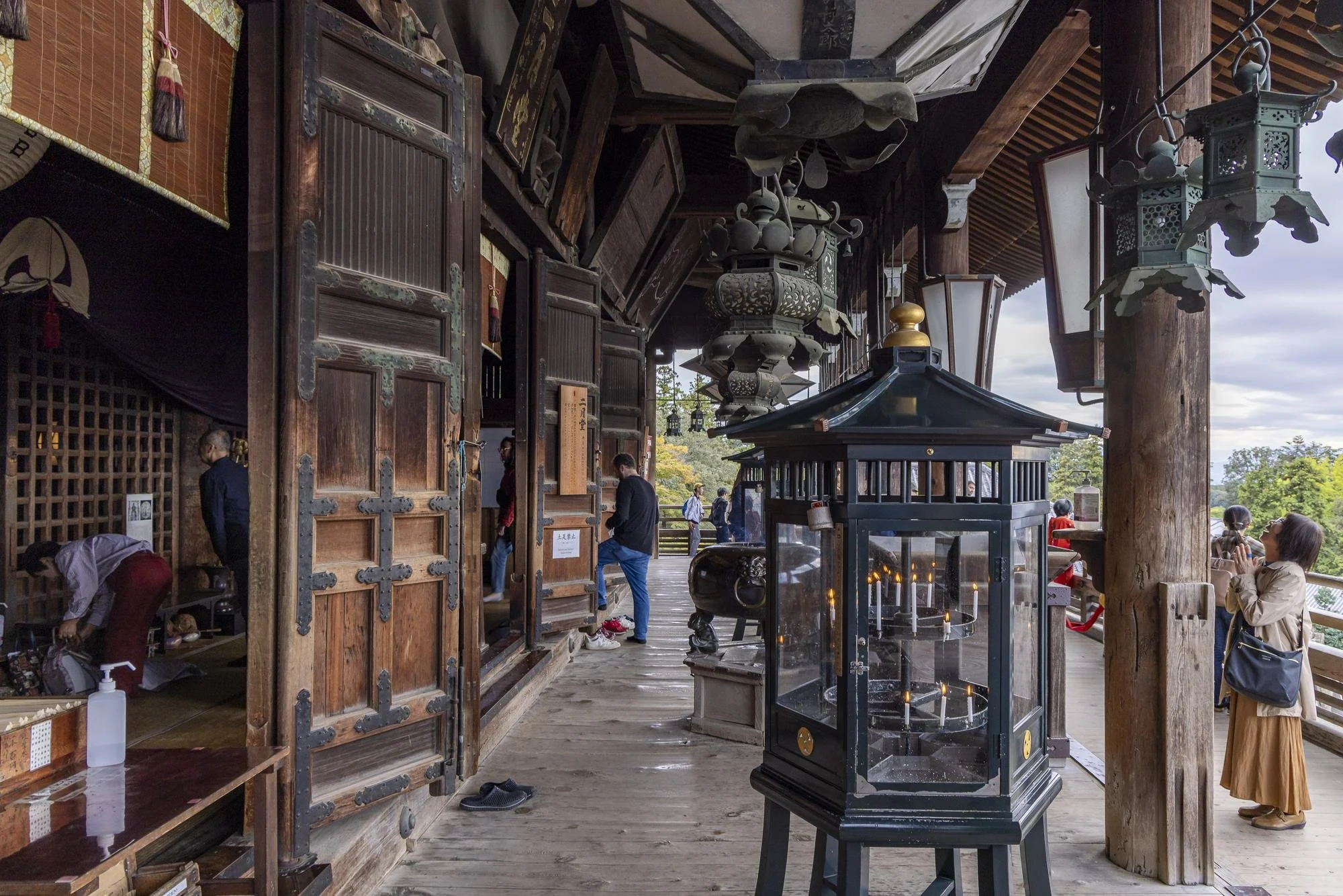 People inside and outside a traditional wooden building, possibly a temple, with hanging lanterns and candles, and a view of trees and sky in the background.